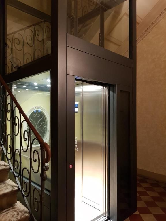 Ascensore con porta aperta accanto alle scale. Struttura in metallo, pareti in vetro e pareti marroni e beige.