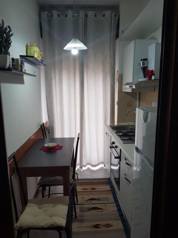 Piccola cucina con tavolo e due sedie. Mobili bianchi e fornelli. Finestra con tende al centro.