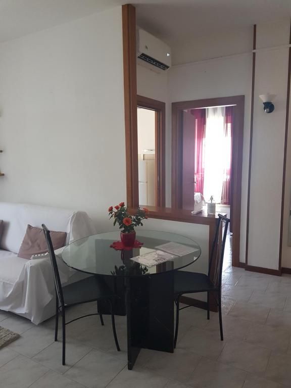 Piccola sala da pranzo con tavolo in vetro, sedie nere e divisorio parziale. Pareti bianche, rose in vaso, porta sullo sfondo.