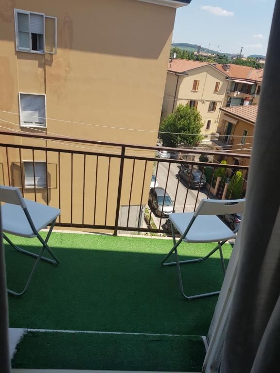 Balcone con due sedie bianche, che si affaccia su una strada con case. Tappeto verde e ringhiera marrone.