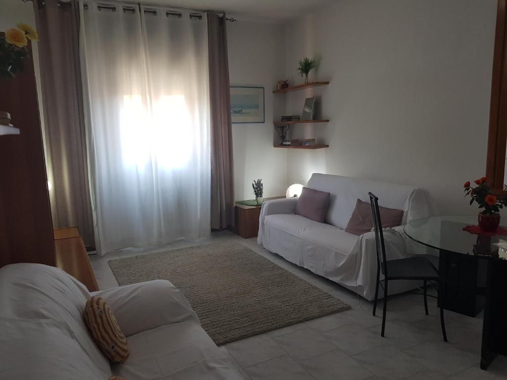 Soggiorno con divani bianchi, finestra con tende, tappeto e scaffali.