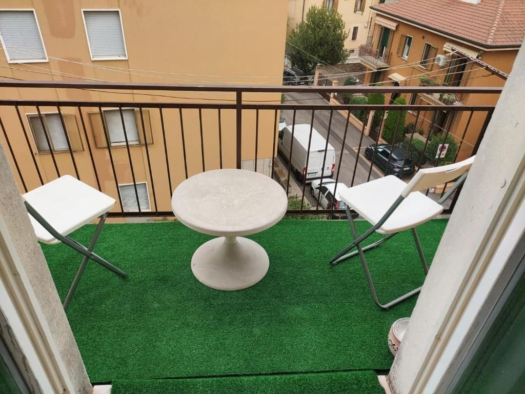 Balcone con due sedie bianche, un tavolino e prato verde artificiale. Vista della strada dall'alto.