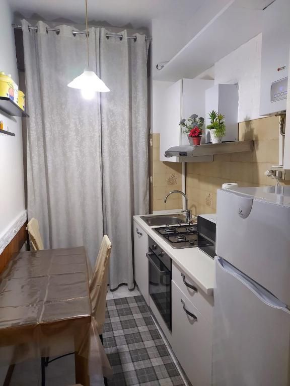 Piccola cucina bianca con fornelli, lavello e frigorifero. Un tavolo e delle sedie si trovano davanti a una finestra con tende.