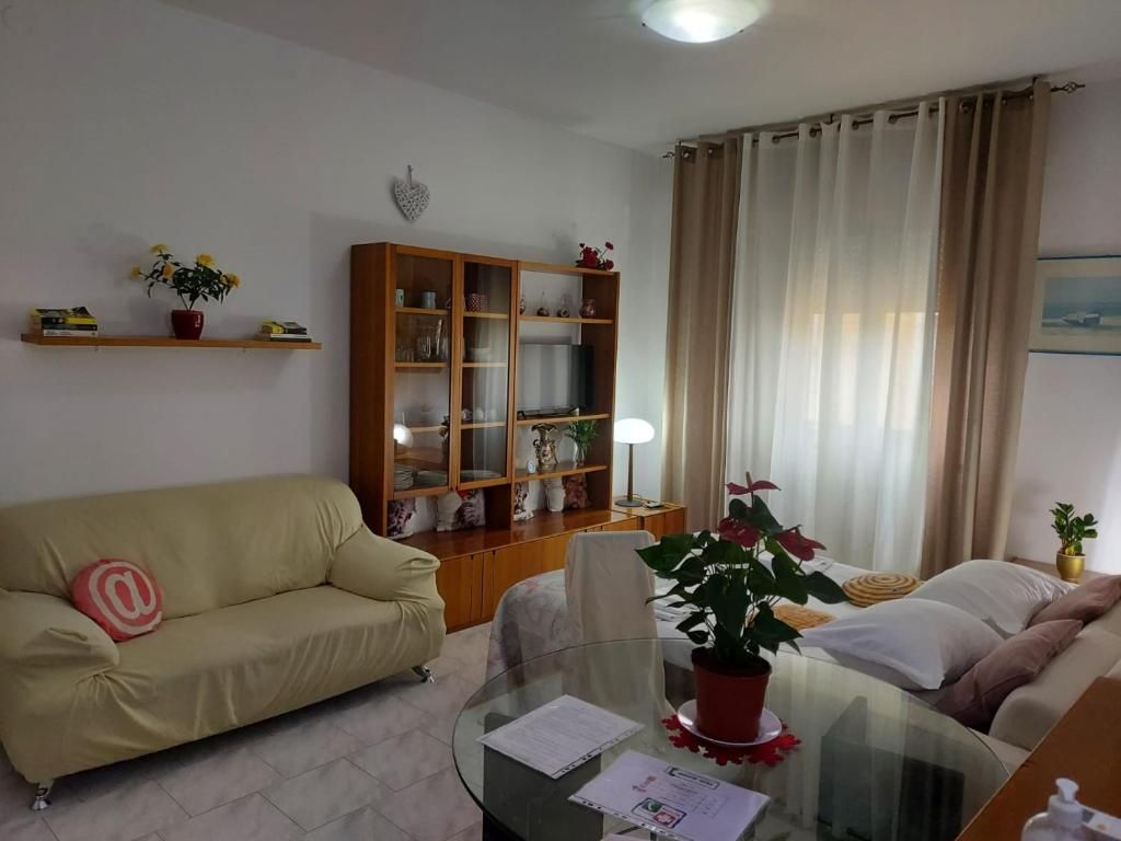 Accogliente soggiorno con divano beige, mobile in legno, tavolo in vetro e tende trasparenti.