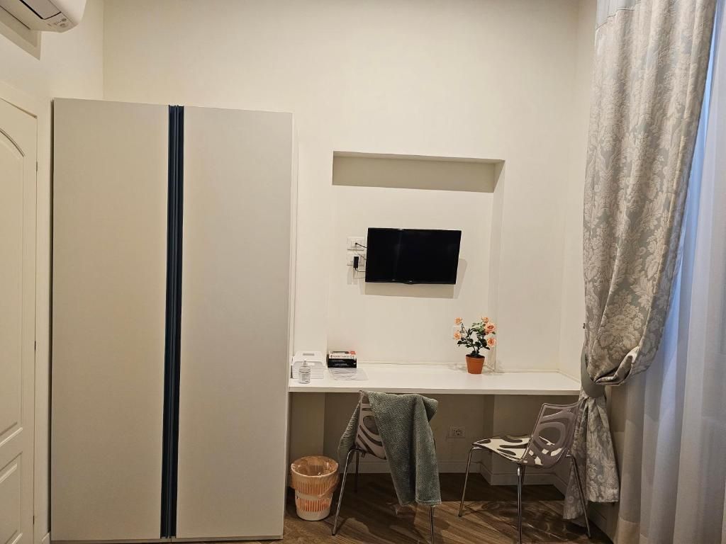 Camera da letto con armadio bianco, scrivania con TV, fiori e finestra con tende trasparenti.