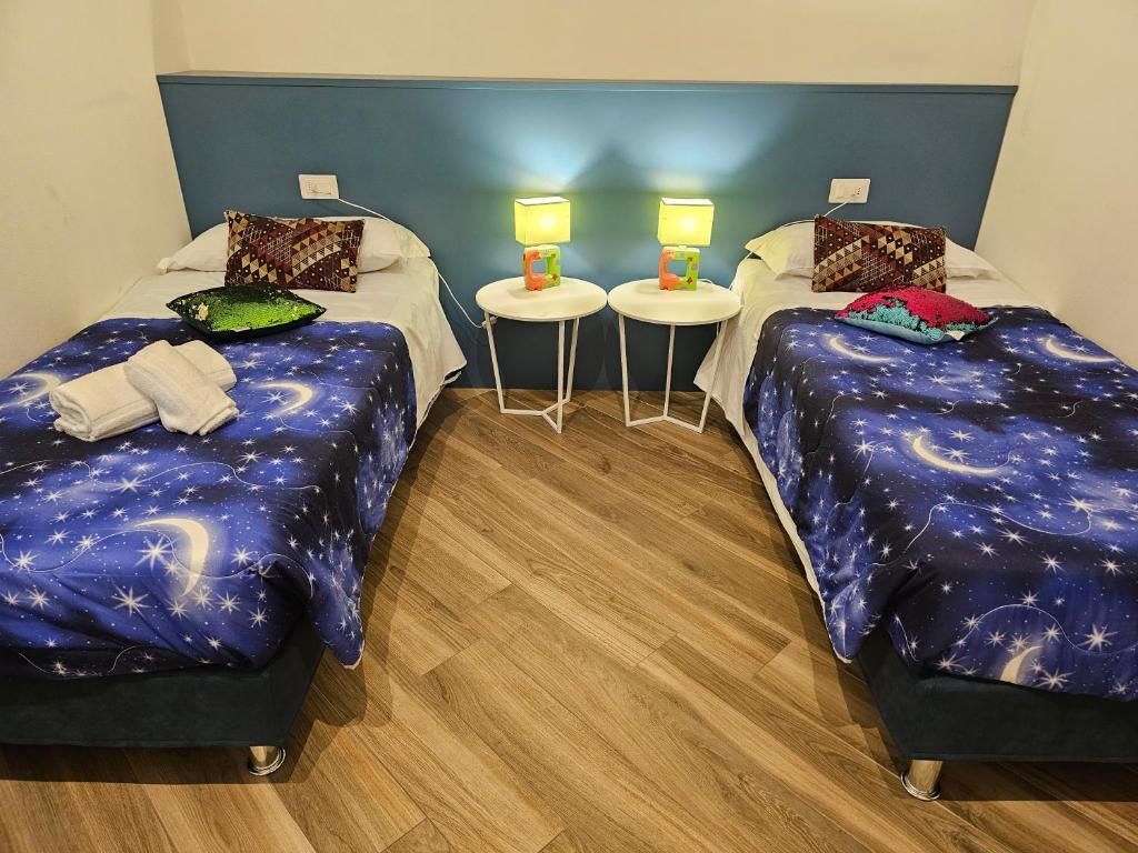 Due letti singoli in una stanza con biancheria da letto a tema galattico. Piccoli tavolini con lampade tra di loro.