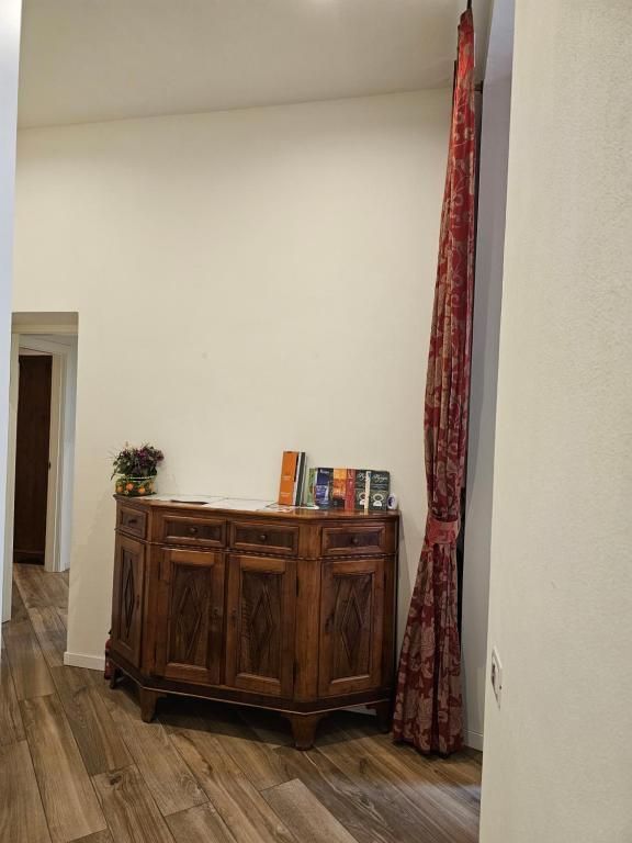 Mobiletto in legno con libri e fiori. Tenda rossa a destra, corridoio con pavimento in legno.