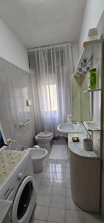 Un bagno piccolo e stretto con lavatrice, WC, bidet e lavandino. Tende trasparenti coprono una finestra.