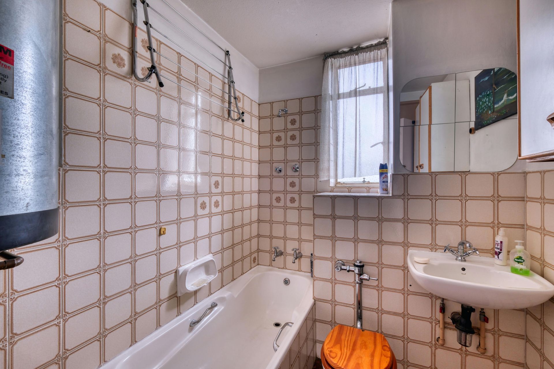 A bathroom with beige tiled walls, a white bathtub, a pedestal sink with a medicine cabinet mirror, and a window.