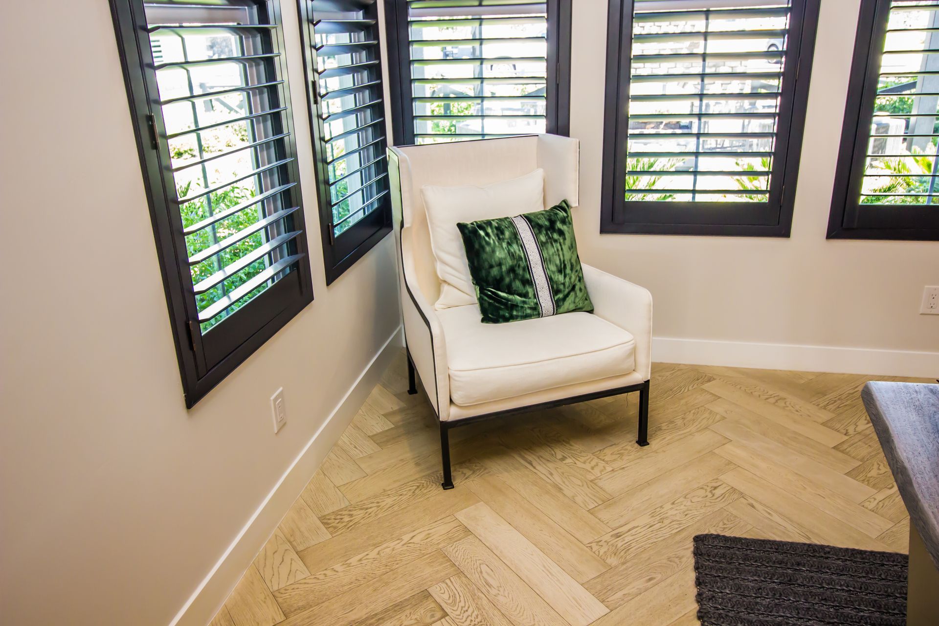 Bright corner room with a white armchair and large shuttered windows over wood flooring.