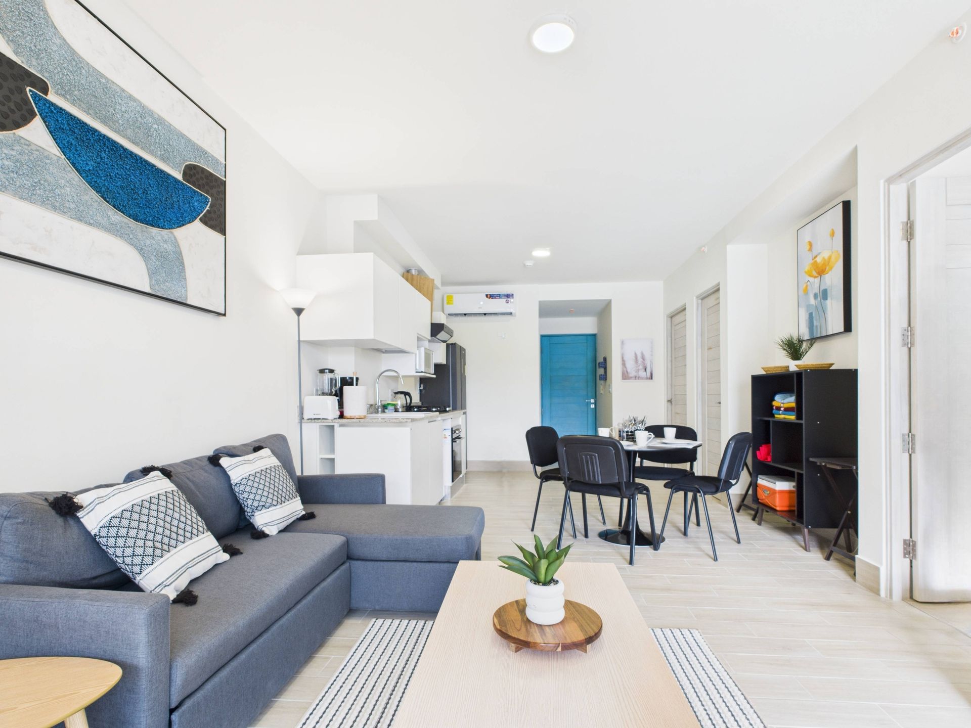 Living room with gray sofa, kitchen, dining area, and artwork. White walls and light wood floors.