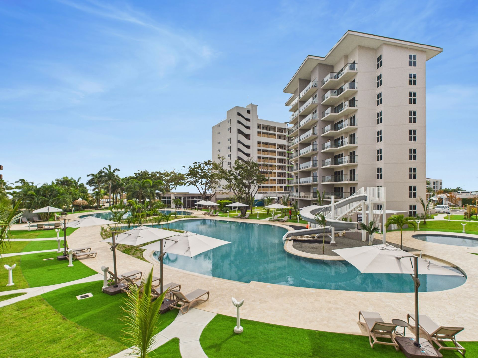 Resort pool with lounge chairs, umbrellas, and multi-story buildings under a blue sky.