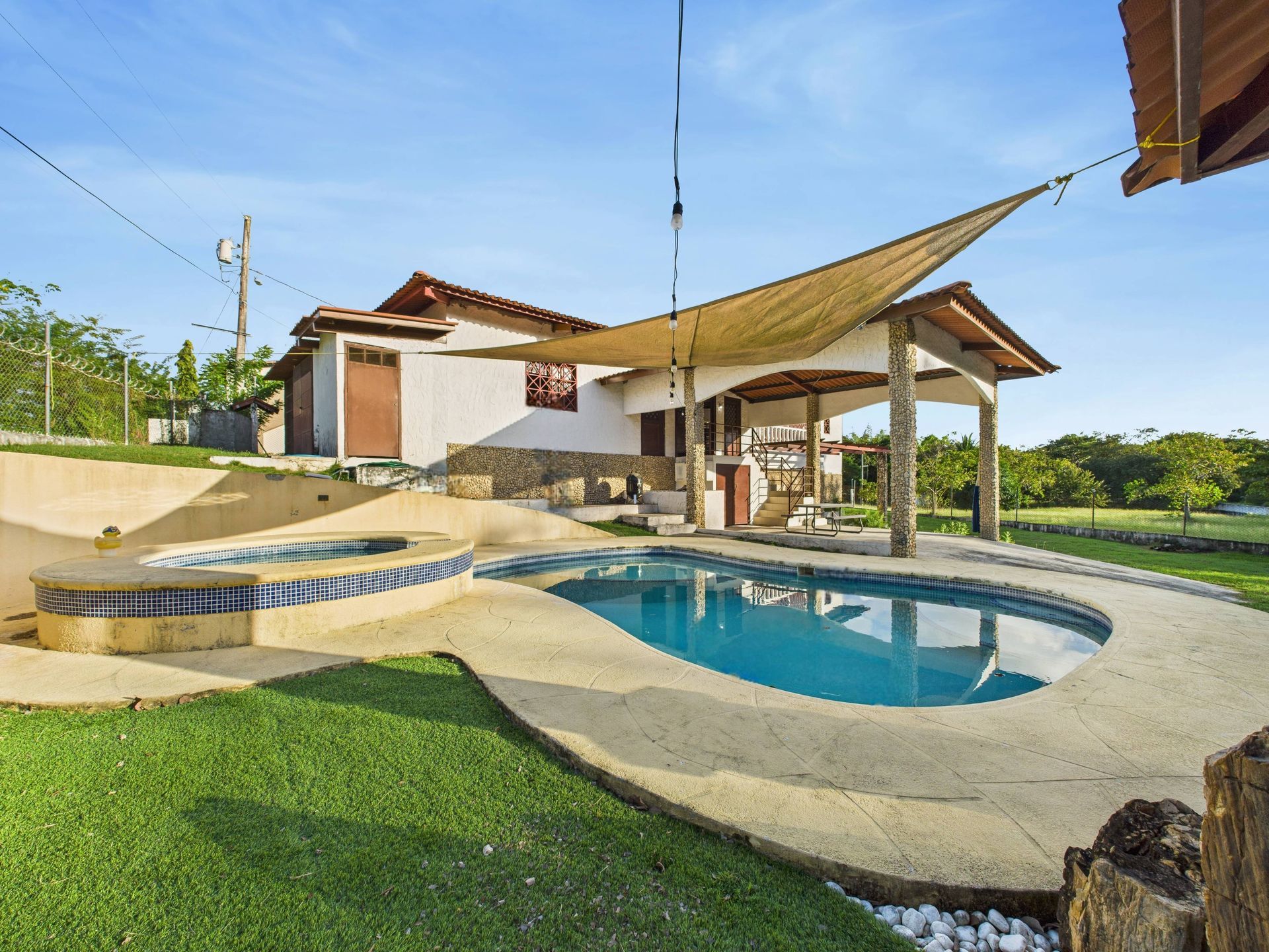 Casa con piscina y patio cubierto. Piscina azul, césped verde, edificio blanco y sombrilla beige.