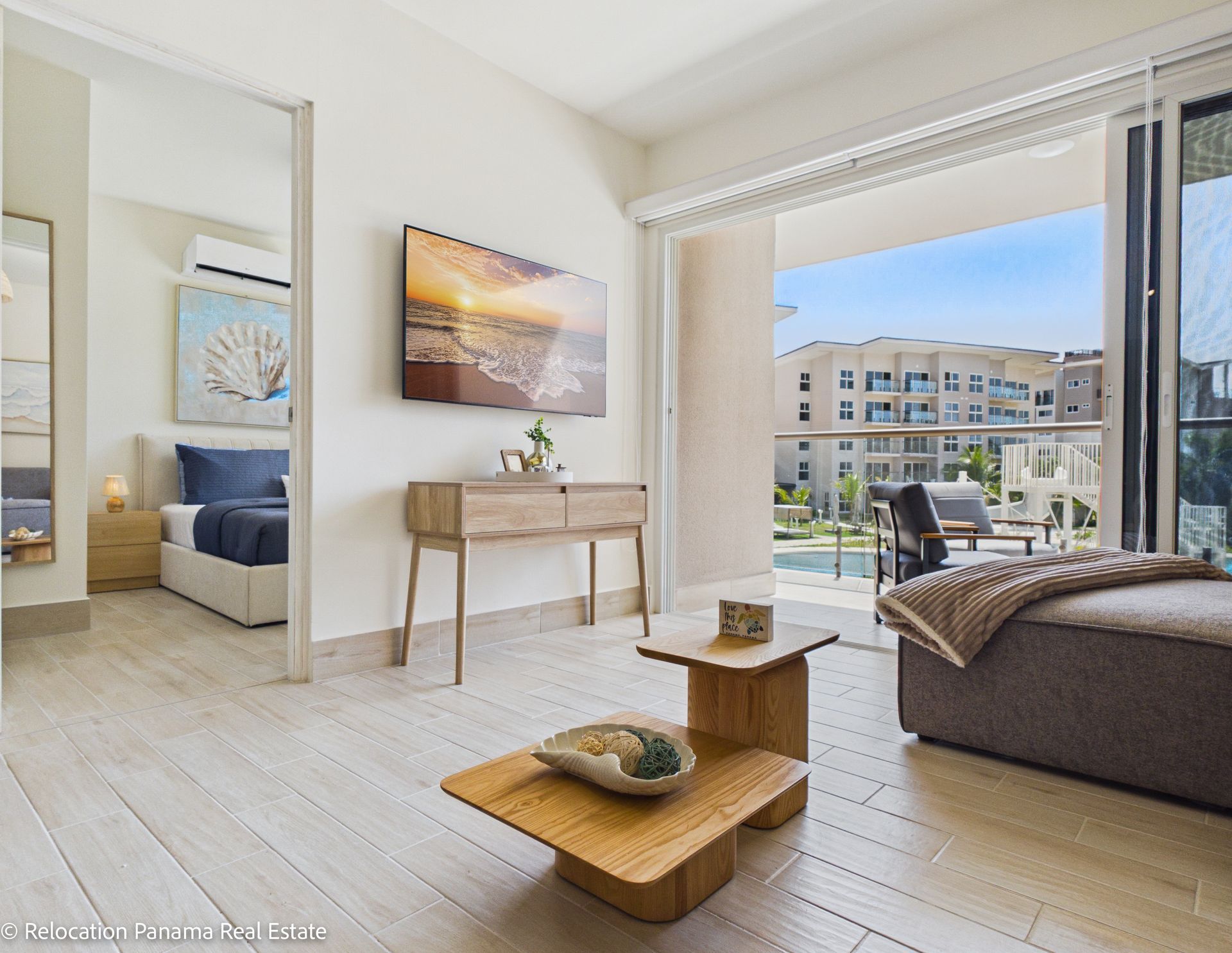 Un luminoso y moderno salón de apartamento con mesa de centro, consola, televisor montado en la pared y balcón con vistas a un complejo.
