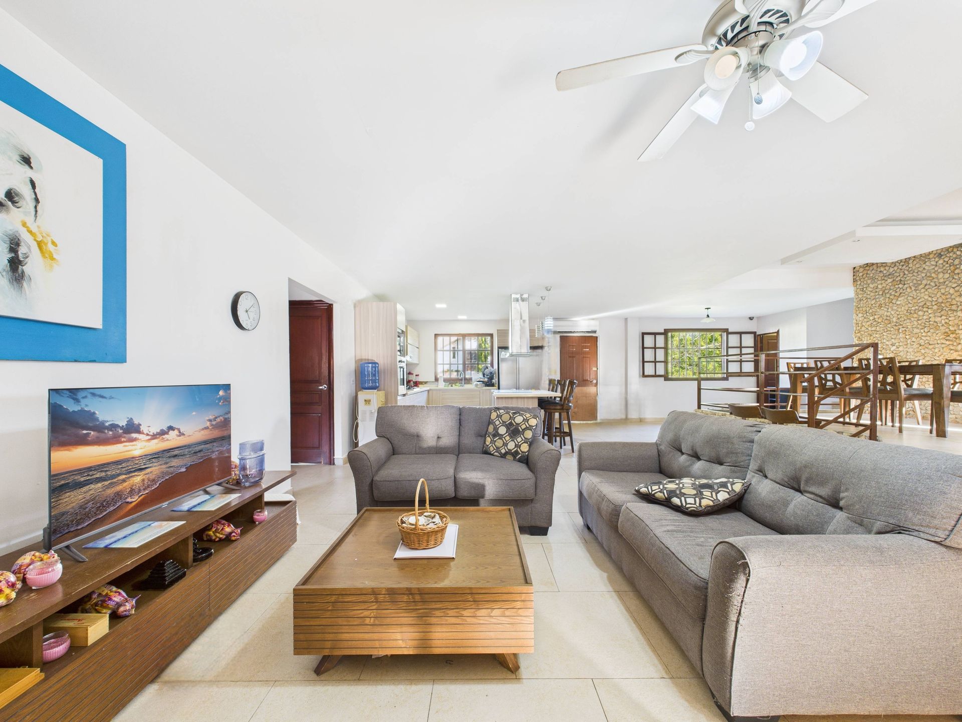 Sala de estar con sofás grises, mesa de centro de madera, TV, abierta a la cocina y al comedor.