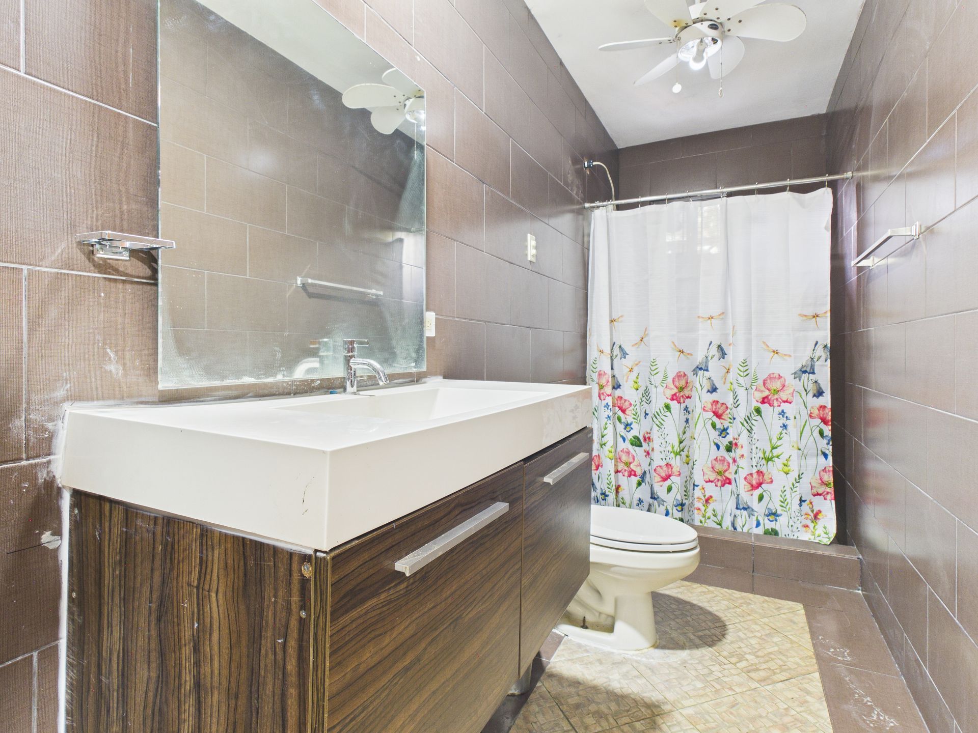 Un baño con paredes revestidas de azulejos marrones, un tocador con acabado de madera, un lavabo blanco, un espejo, un inodoro y una cortina de ducha con estampado floral.