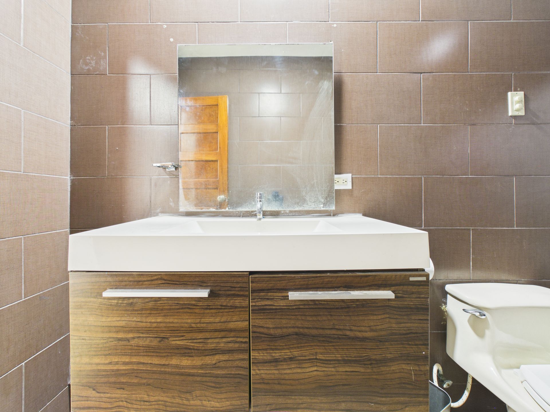 Un tocador de baño con encimera blanca y armarios con acabado de madera, centrado bajo un espejo cuadrado sobre paredes alicatadas.