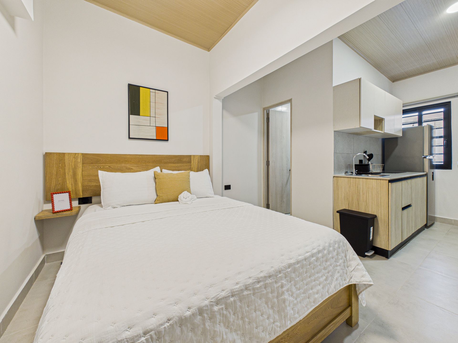 Un estudio con una cama junto a un cabecero revestido de madera, al lado de una pequeña zona de cocina con nevera en Bethania, fotografía inmobiliaria.