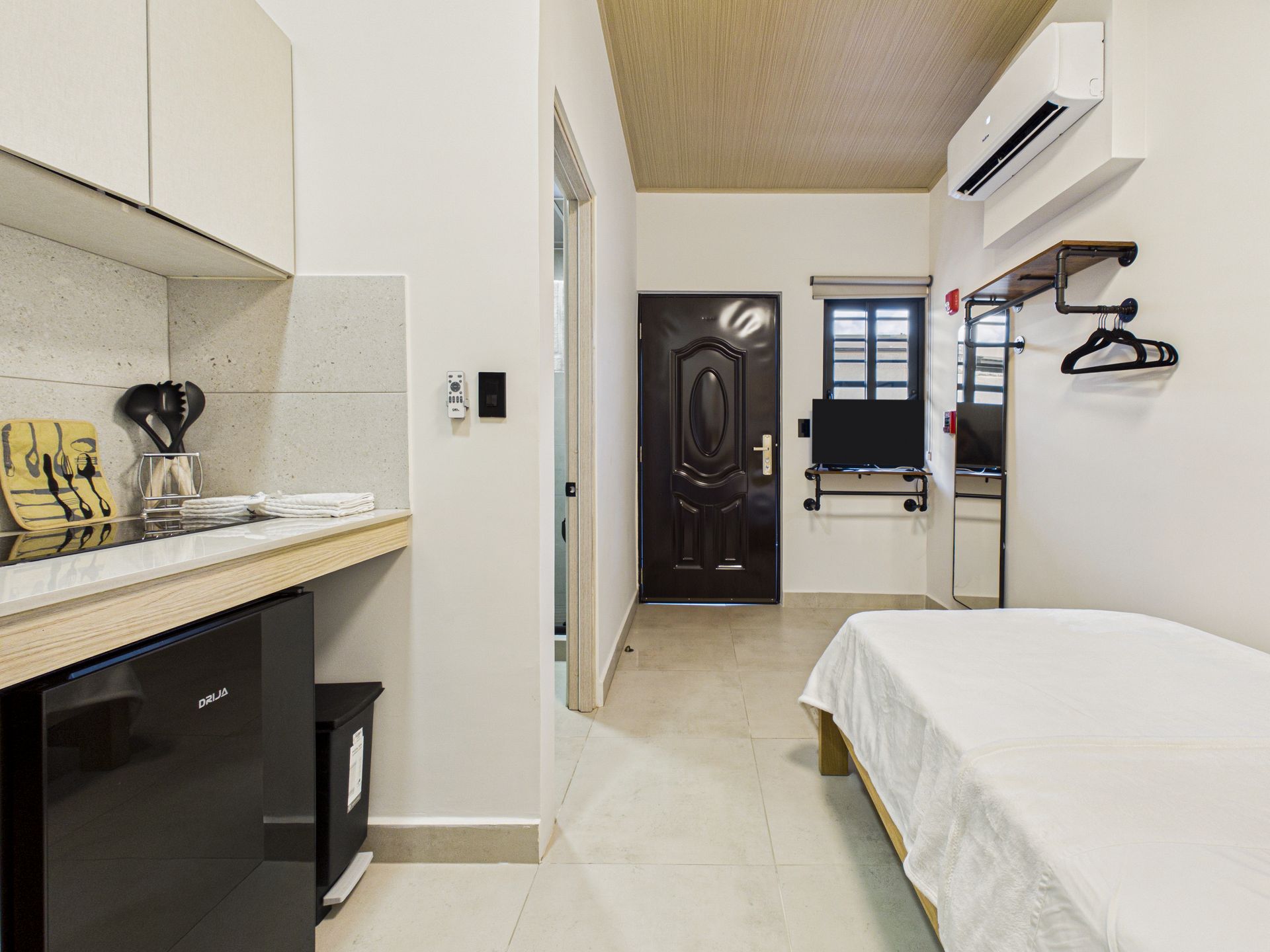 Un pequeño y ordenado estudio con zona de cocina, una cama individual, puerta negra y aire acondicionado de pared. en Bethania, fotografía inmobiliaria.