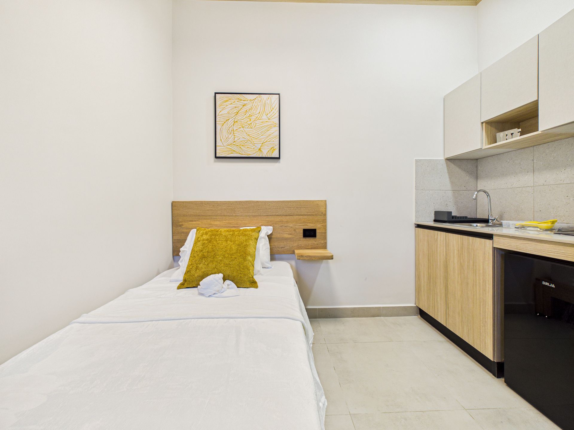 Un estudio minimalista con una cama individual con almohada amarilla, una pequeña mesita de noche de madera y una cocina compacta. en Bethania, fotografía inmobiliaria.