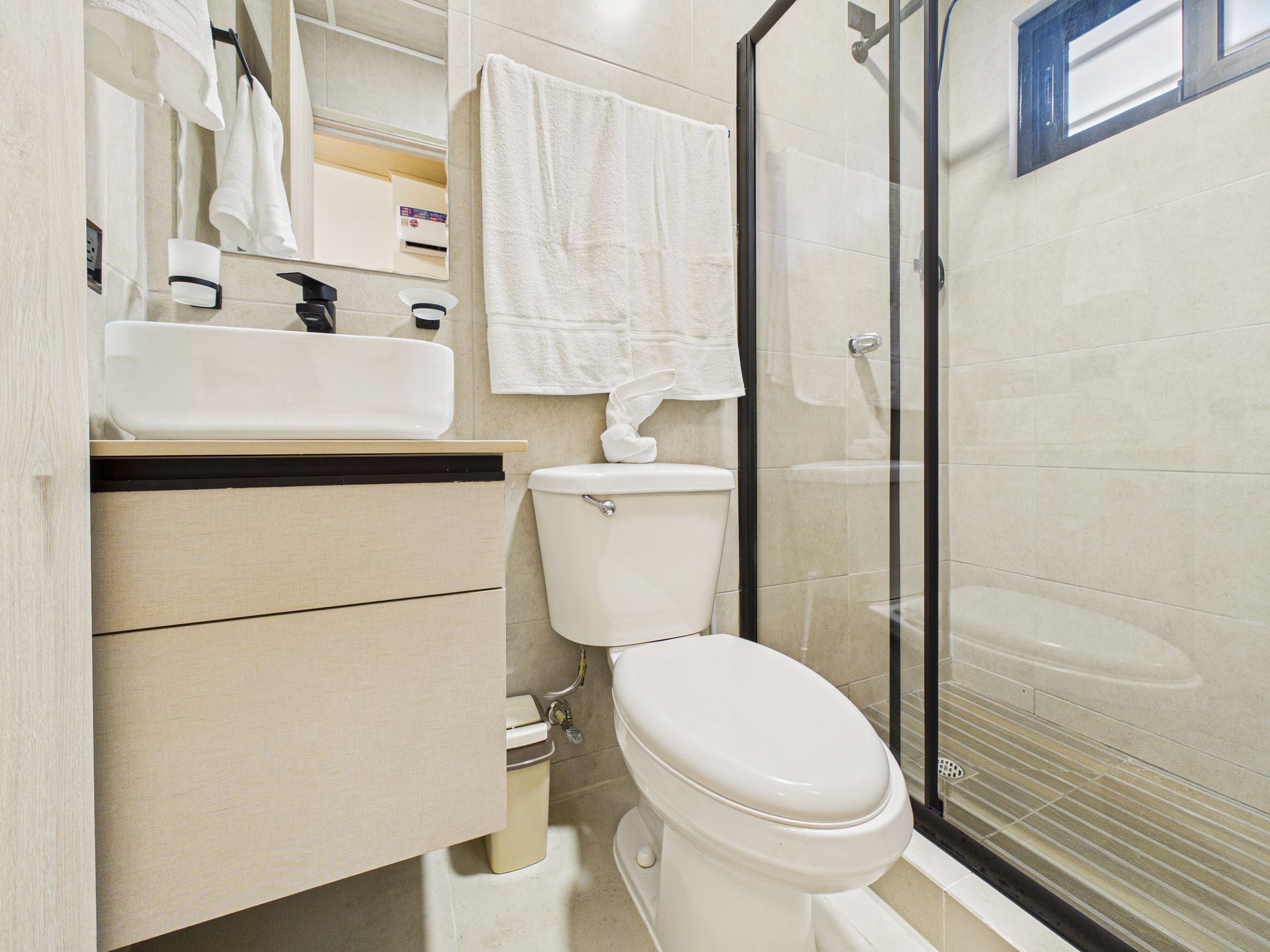Un baño compacto con un tocador blanco con lavabo moderno, un inodoro blanco y una ducha con mampara de cristal.en Bethania, fotografía inmobiliaria.
