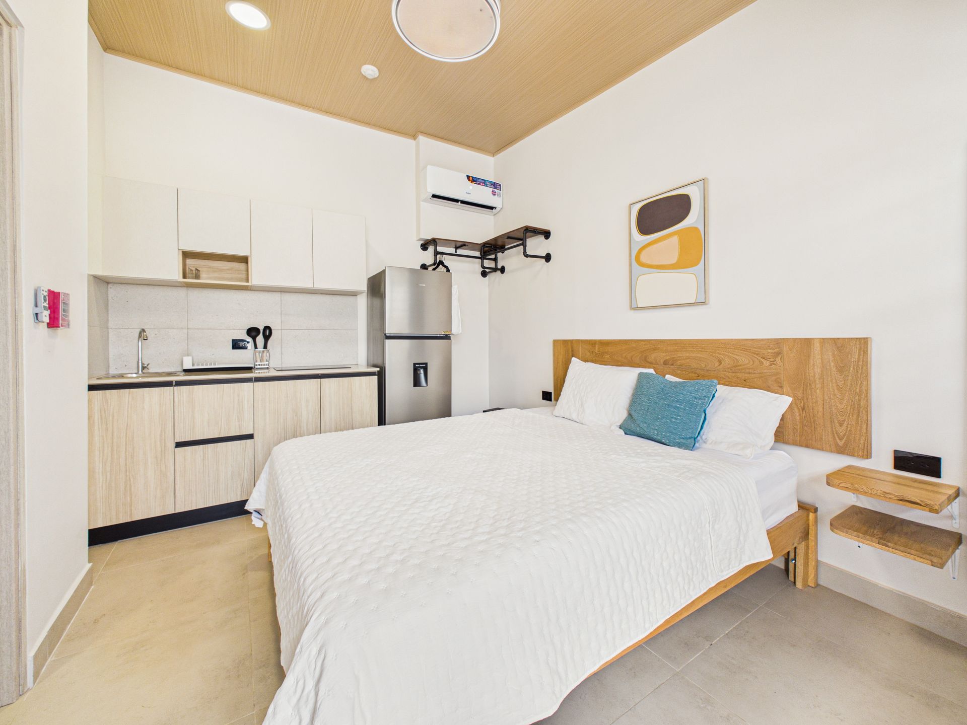 Un pequeño estudio con cocina, una cama con ropa de cama blanca y una almohada azul, y detalles en madera clara. 