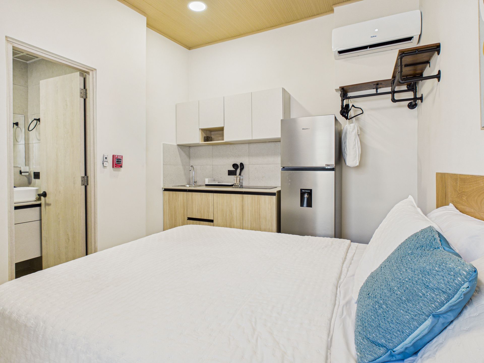 El interior de una habitación de hotel cuenta con una cama con almohada azul, una cocina pequeña con nevera de acero inoxidable y una puerta de baño. en Bethania, fotografía inmobiliaria.