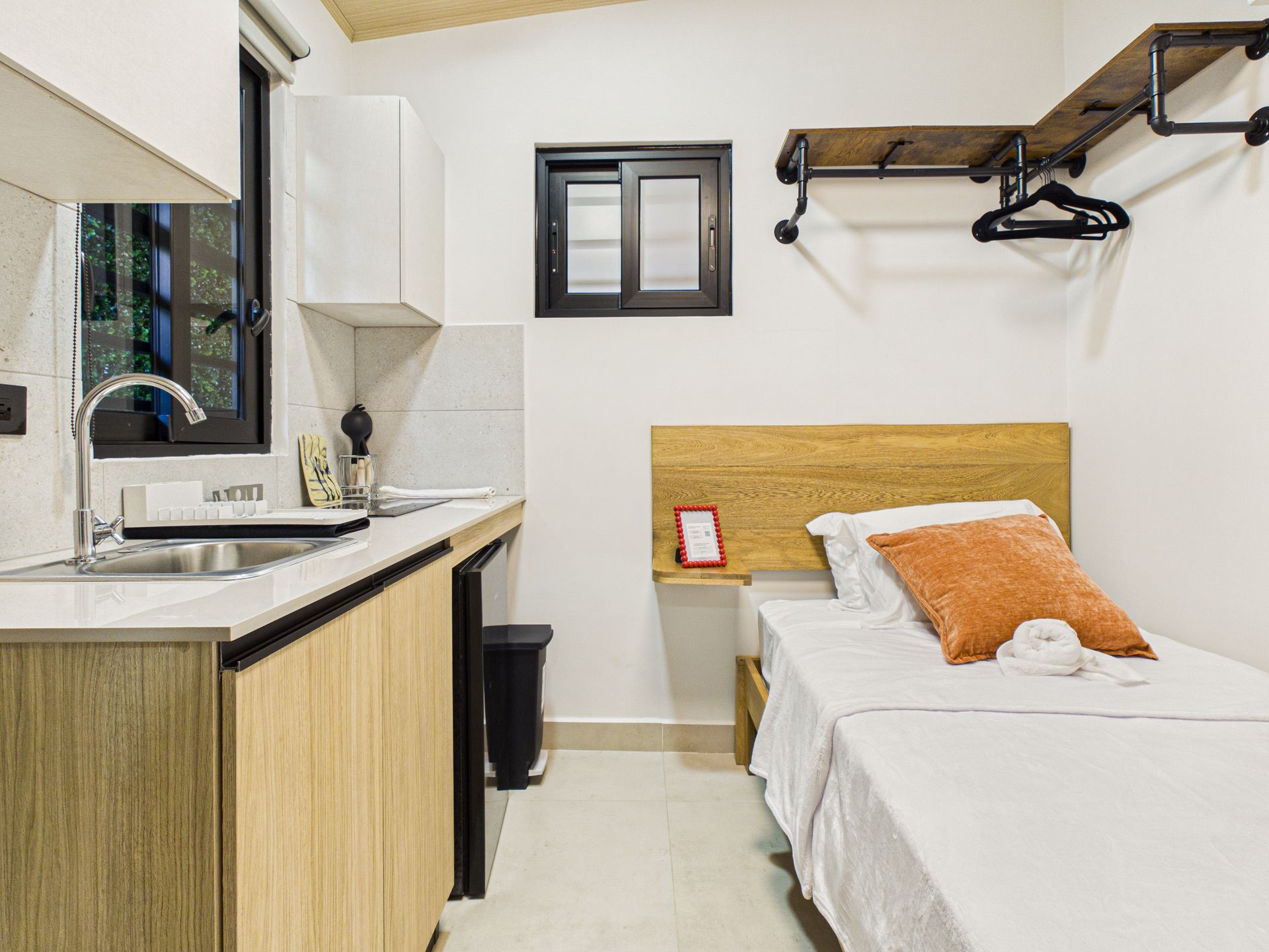 Un estudio compacto que cuenta con una cocina americana con armarios de madera, un fregadero y una cama individual con una almohada naranja. en Bethania, Ciudad de Panamá. Fotografía de real estate