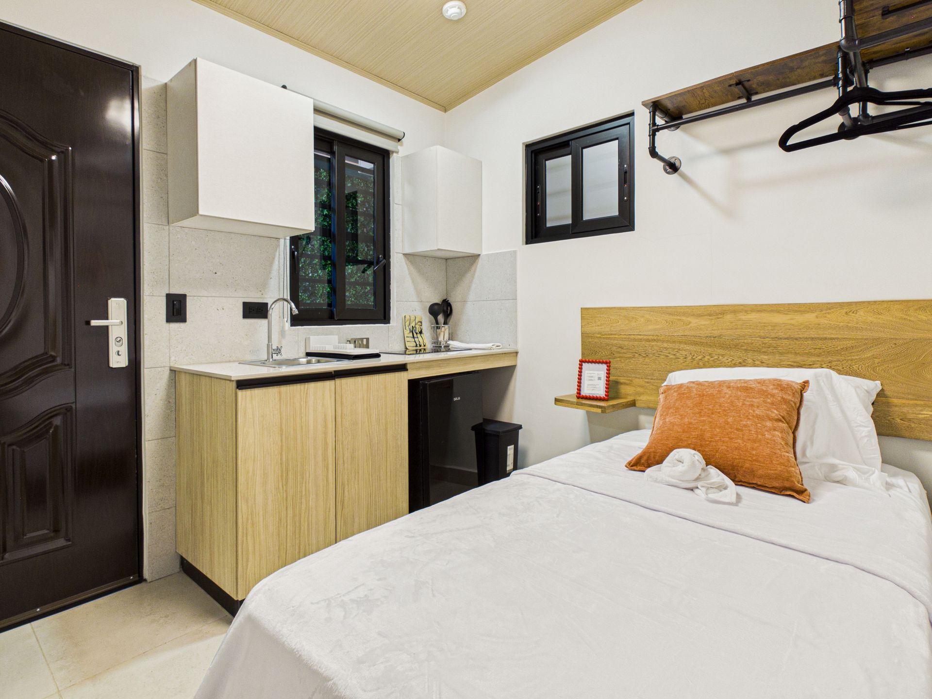 Un estudio compacto con una cama individual con almohada naranja, una cocina americana con armarios de madera y una puerta oscura. en Bethania, Ciudad de Panamá. Fotografía de real estate