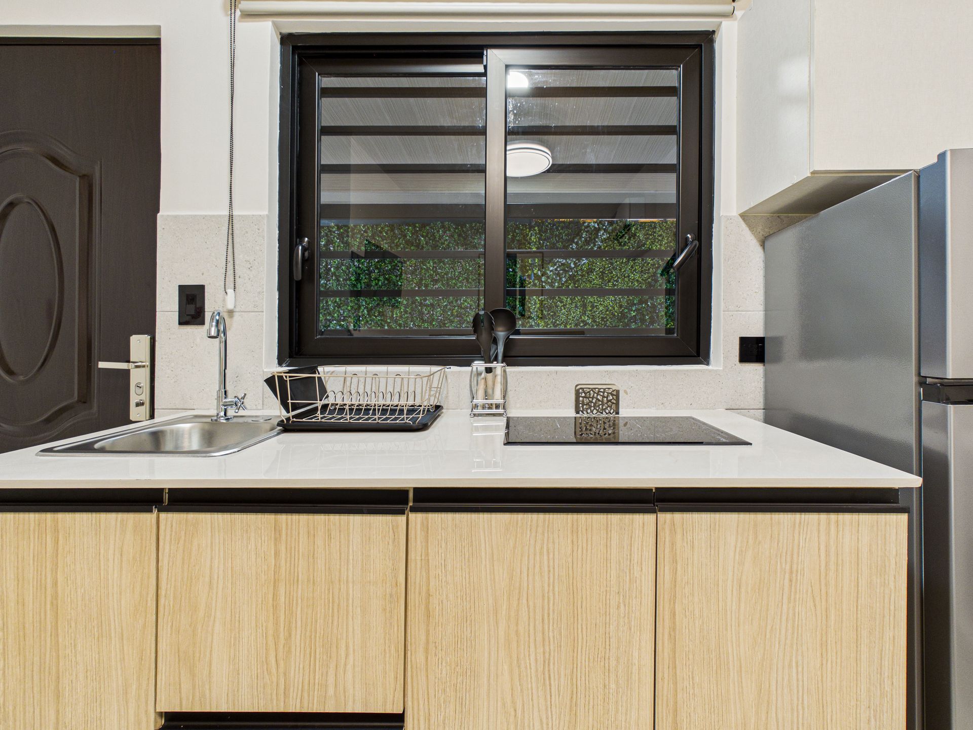 Una moderna encimera de cocina con fregadero, placa de inducción y armarios de madera clara debajo de una ventana con marco oscuro. en Bethania, Ciudad de Panamá. Fotografía de real estate