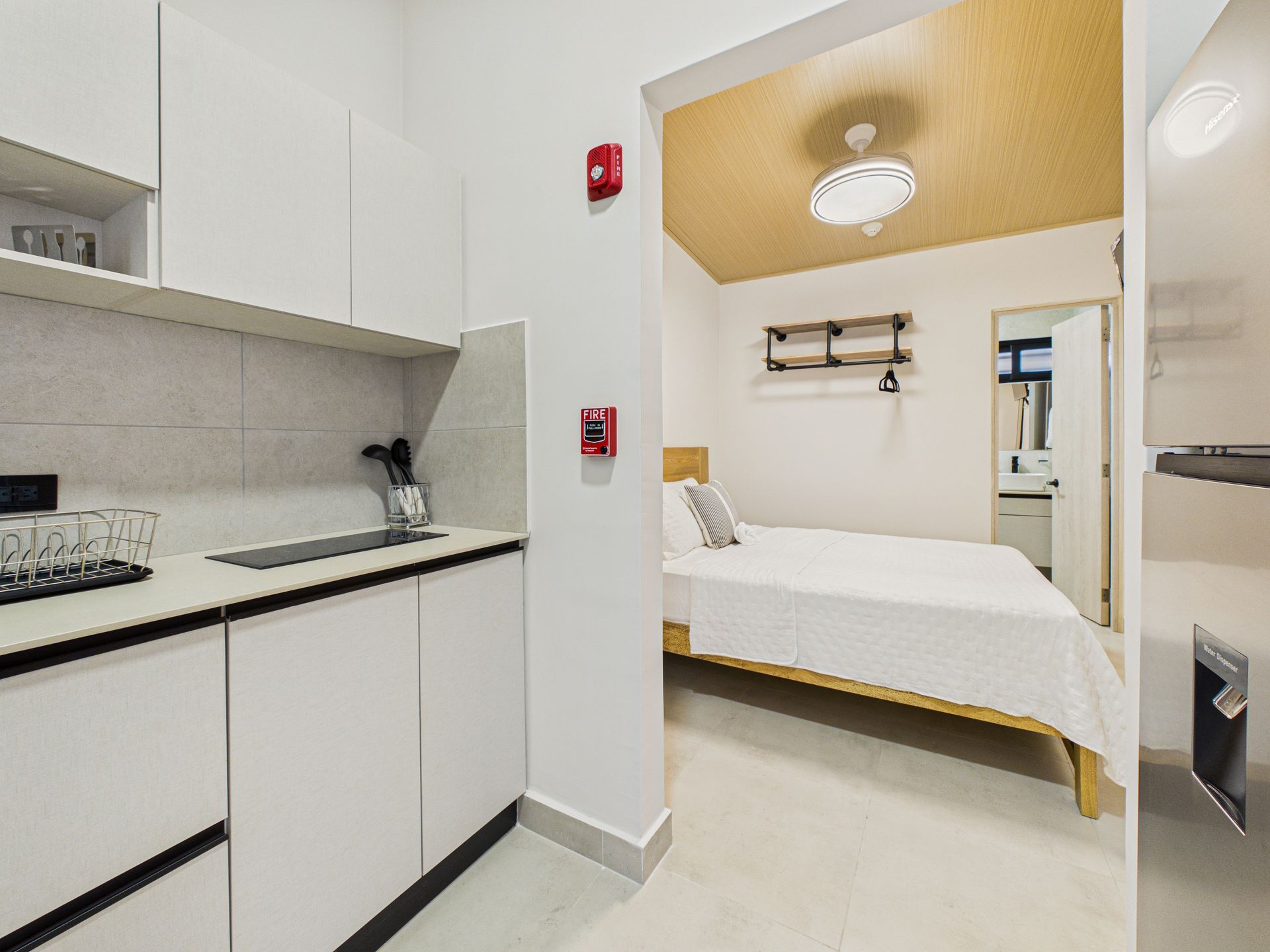 Una pequeña cocina con armarios blancos y encimera oscura, que da paso a un dormitorio con cama blanca y techo de madera clara. en Bethania, Ciudad de Panamá. Fotografía de real estate