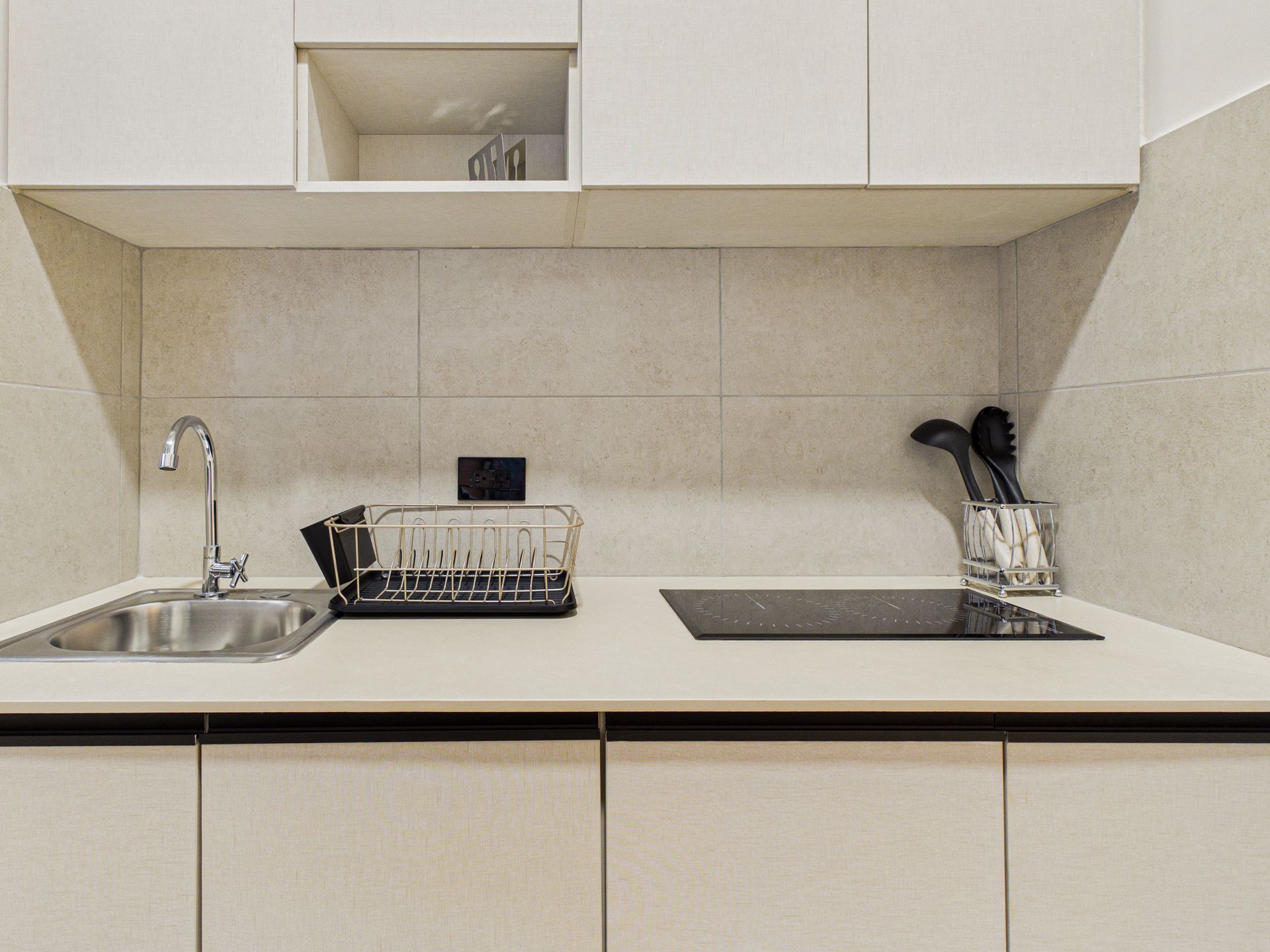 Una encimera de cocina minimalista con fregadero, escurridor de platos, placa eléctrica y soporte para utensilios sobre azulejos de color neutro. 