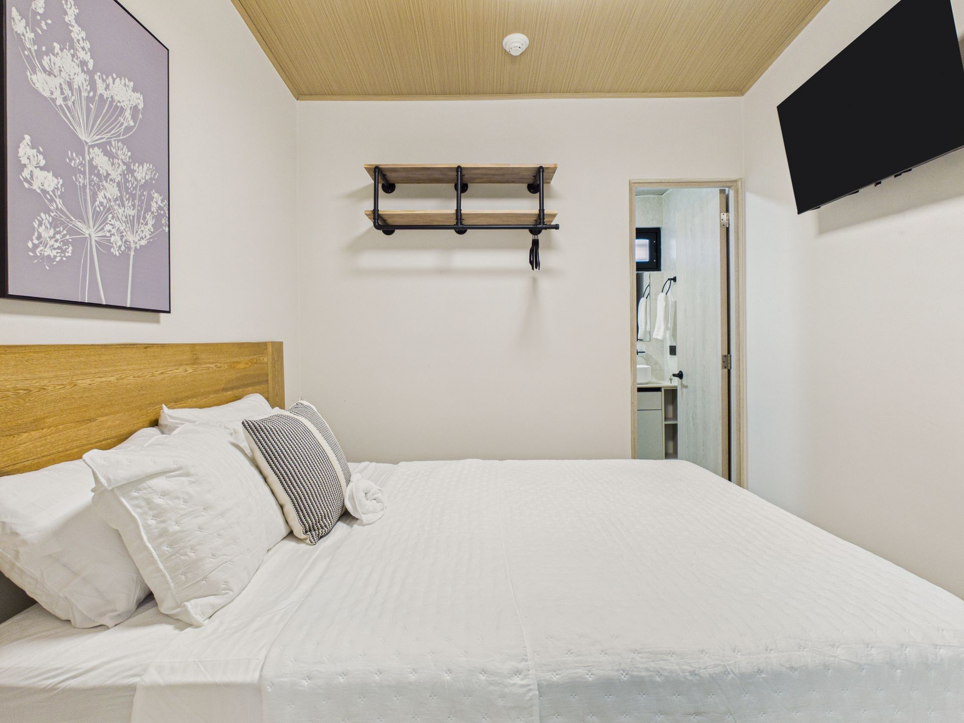 Una habitación de hotel luminosa y moderna con cama con sábanas blancas, cabecero de madera, estantes en la pared y televisor de pantalla plana. en Bethania, Ciudad de Panamá. Fotografía de real estate