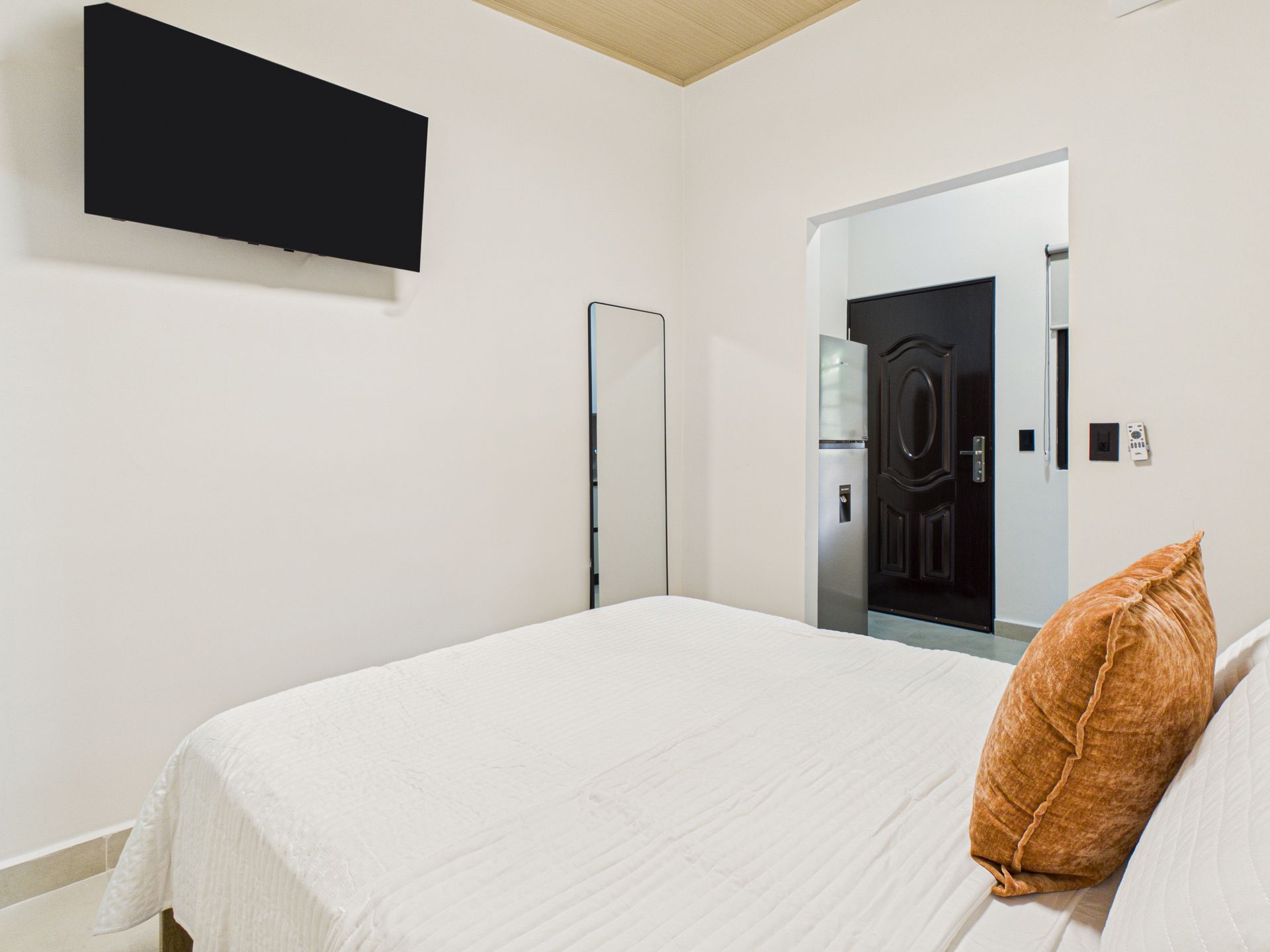 Un dormitorio minimalista con una cama, ropa de cama blanca, un cojín decorativo naranja, un televisor montado en la pared y una puerta que da a un pasillo. en Bethania, Ciudad de Panamá. Fotografía de real estate
