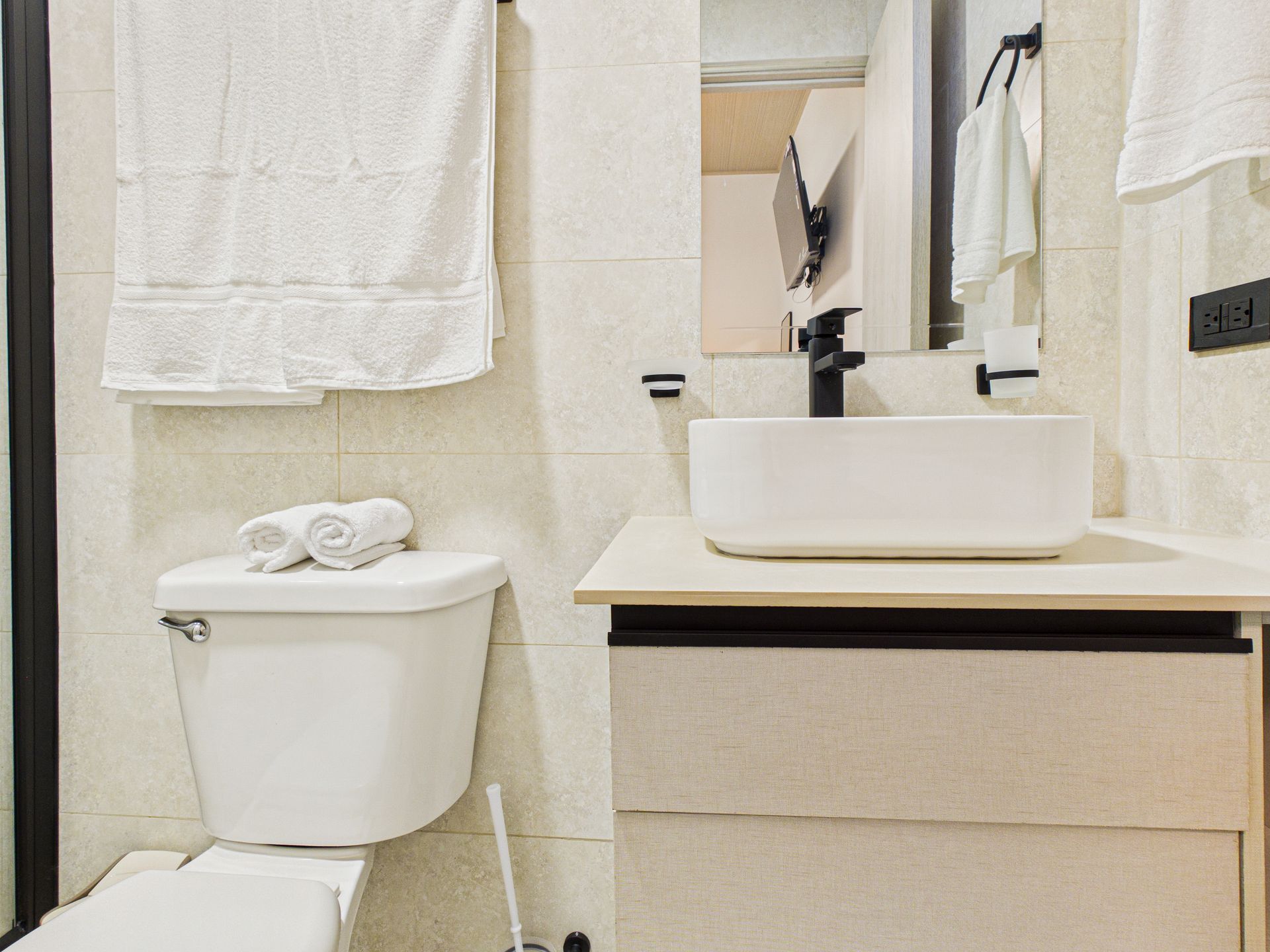 Un baño moderno en tonos neutros que cuenta con inodoro blanco, lavabo sobre encimera, tocador y toallas blancas colgantes. en Bethania, Ciudad de Panamá. Fotografía de real estate