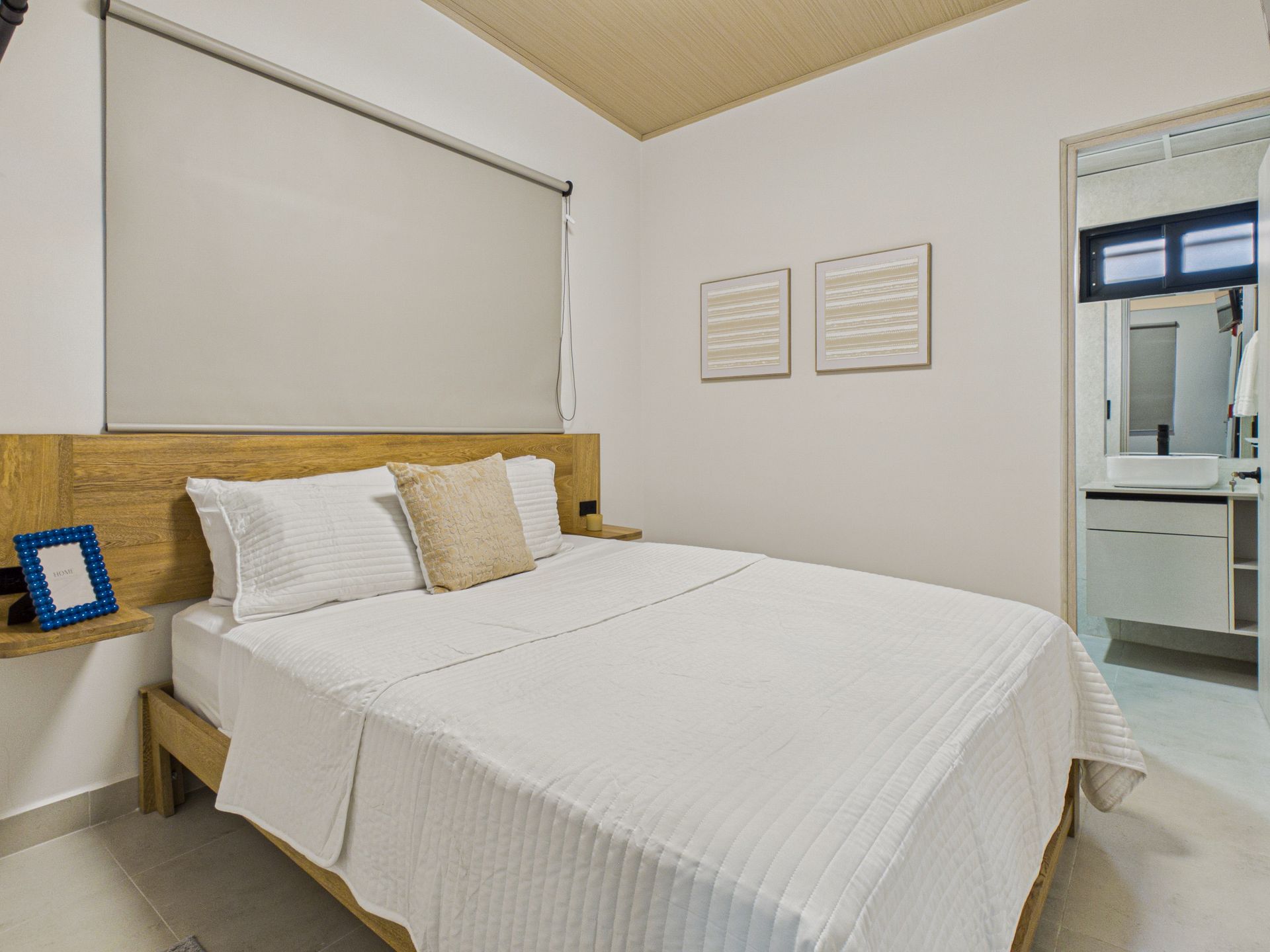 Un dormitorio con una cama de matrimonio con ropa de cama blanca, un cabecero de madera clara, dos cuadros en la pared y un baño privado abierto. en Bethania, Ciudad de Panamá. Fotografía de real estate