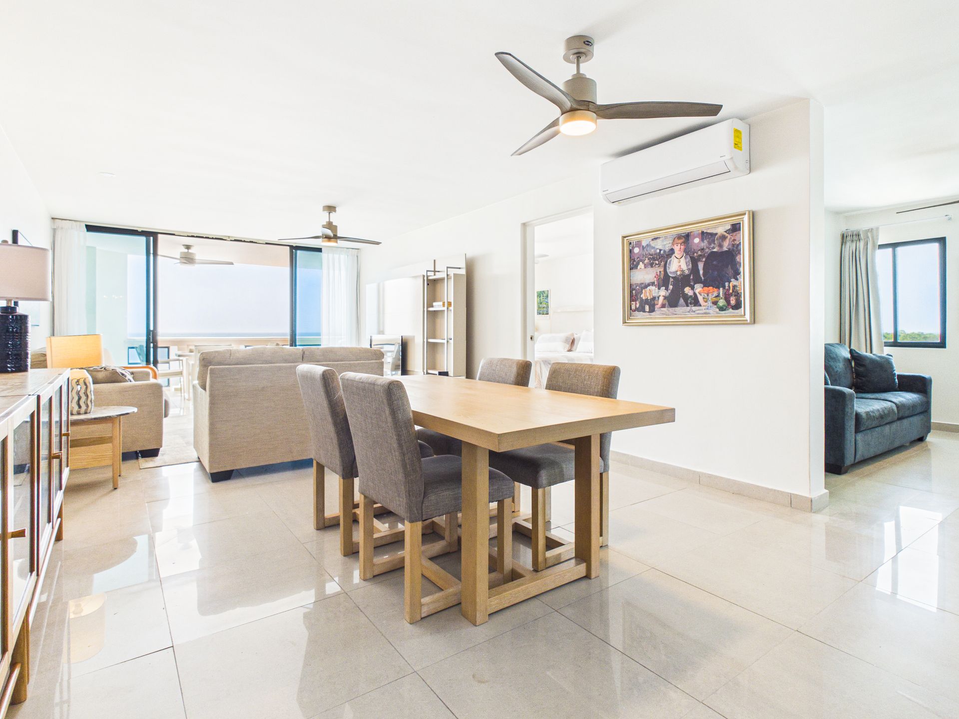 Bright open-plan living room with dining table, sofa, ceiling fan, and large windows overlooking the balcony