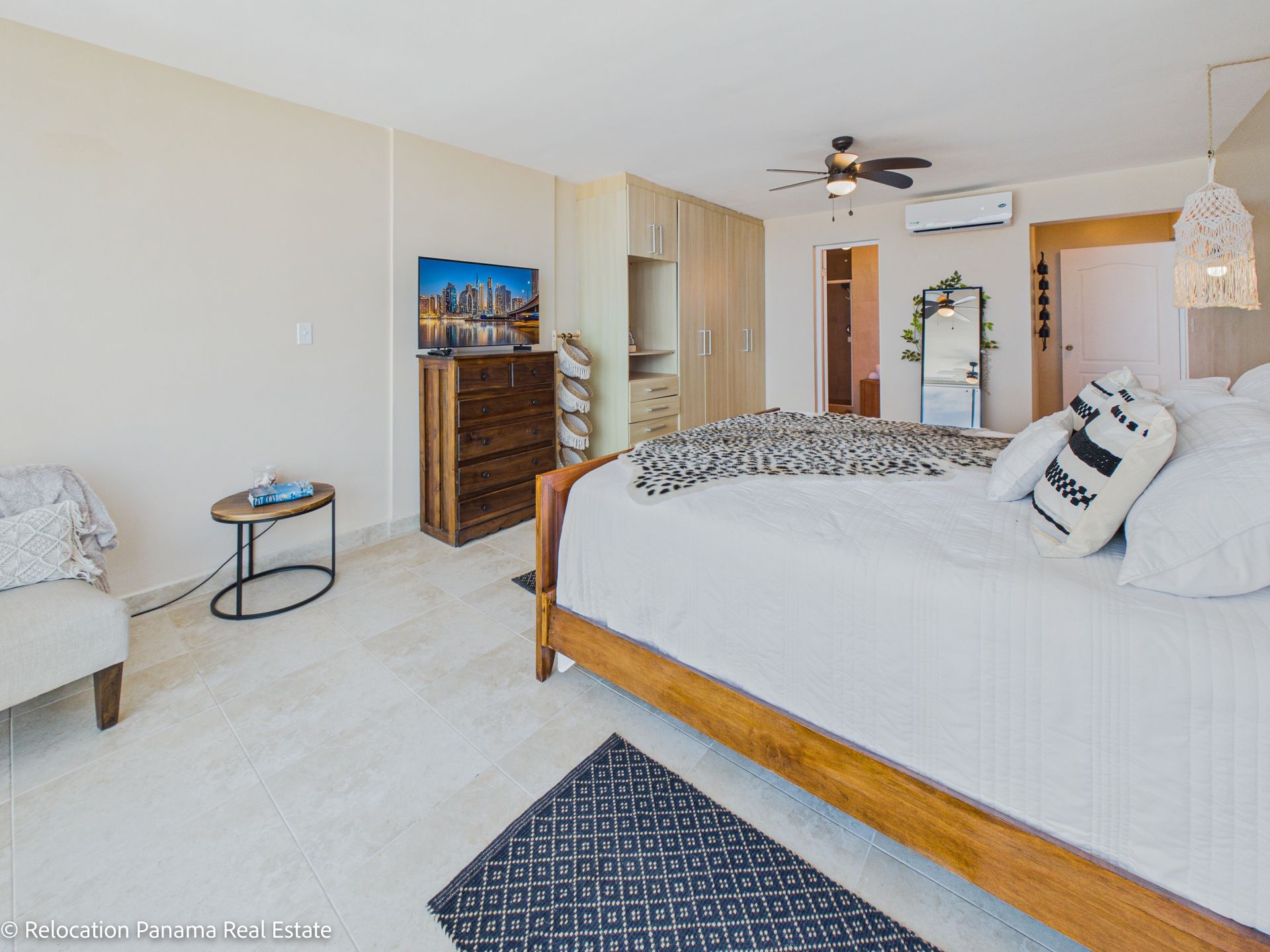 Un dormitorio moderno y luminoso cuenta con una cama con ropa de cama blanca, un marco de madera, una alfombra estampada y un televisor sobre una cómoda.
