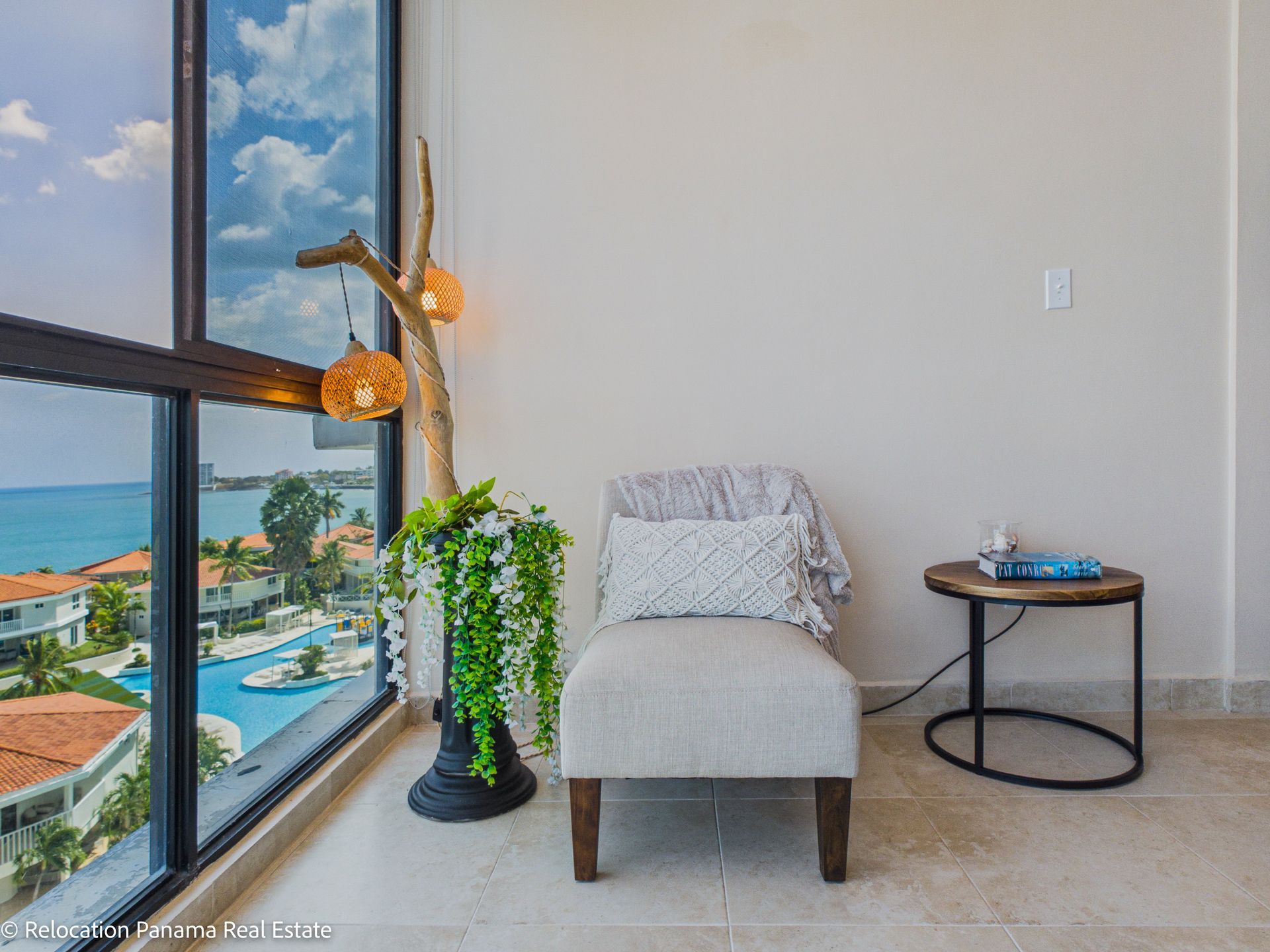 Una acogedora zona de estar junto a una ventana con vistas a un complejo turístico de playa, que cuenta con una silla clara, una mesa auxiliar y una lámpara decorativa.