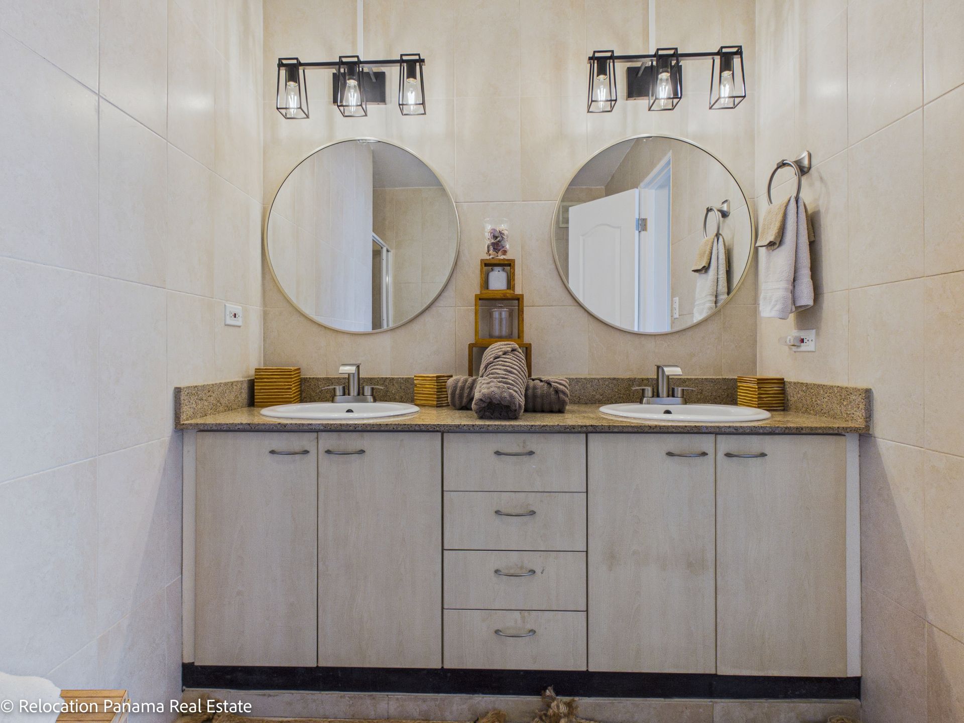 Un moderno mueble de baño con dos lavabos, espejos circulares, elementos decorativos apilados y armarios de madera clara sobre azulejos beige.