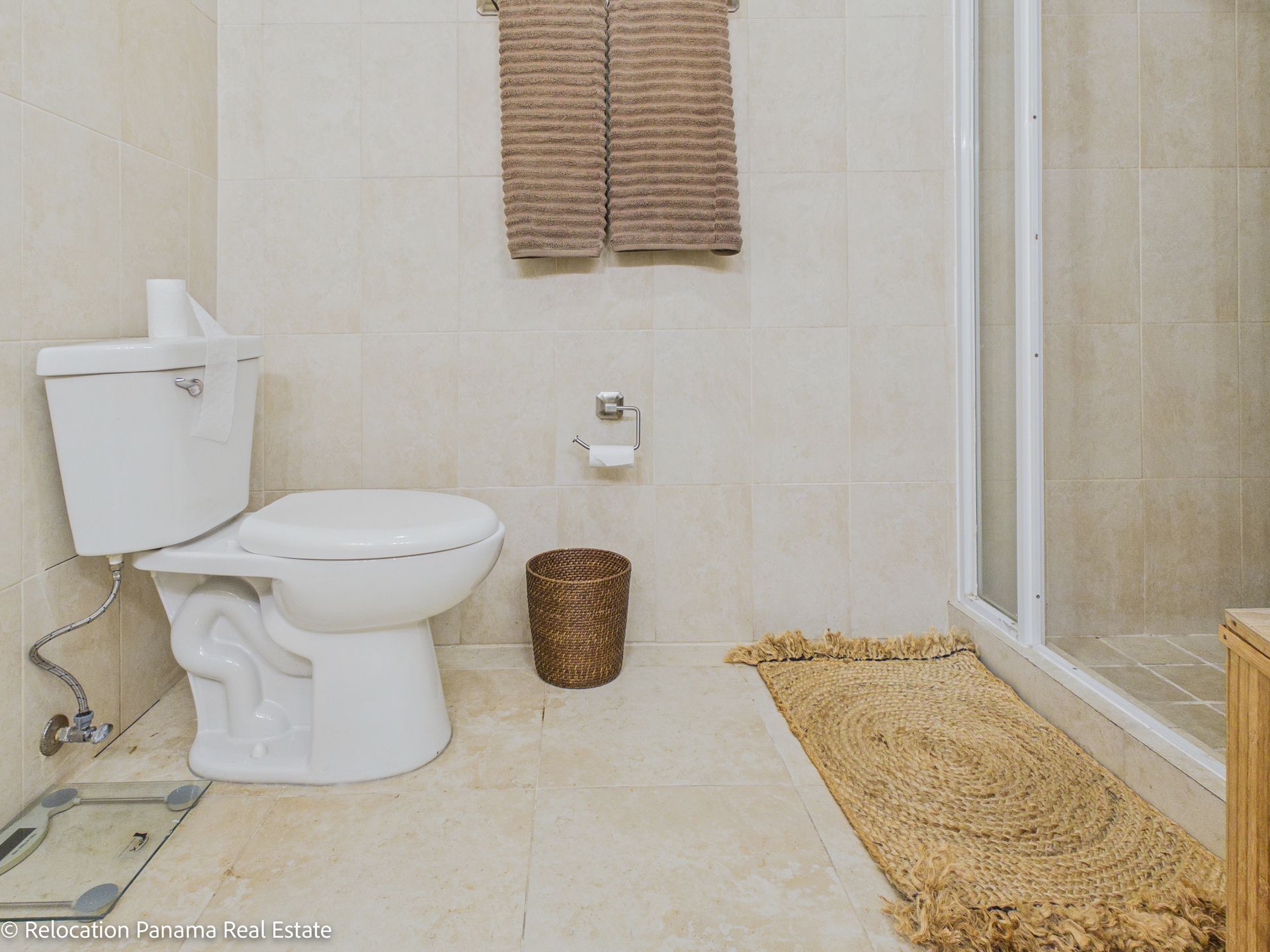 Un baño con inodoro blanco, papelera marrón, toallas colgadas y una alfombra texturizada cerca de la ducha.