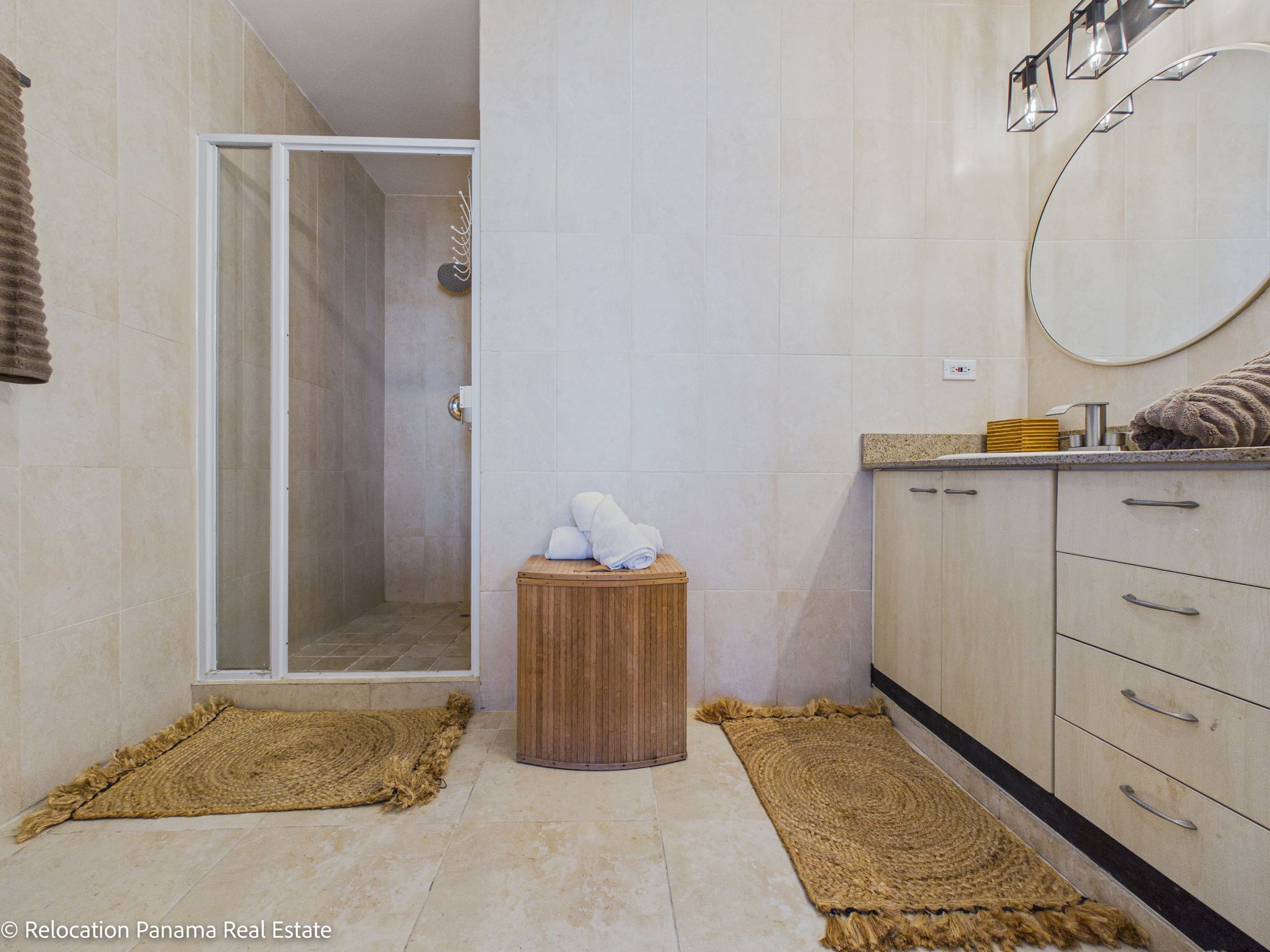 El baño moderno cuenta con una ducha a ras de suelo con puerta de cristal, un tocador de madera con espejo redondo y dos alfombras de yute texturizado.