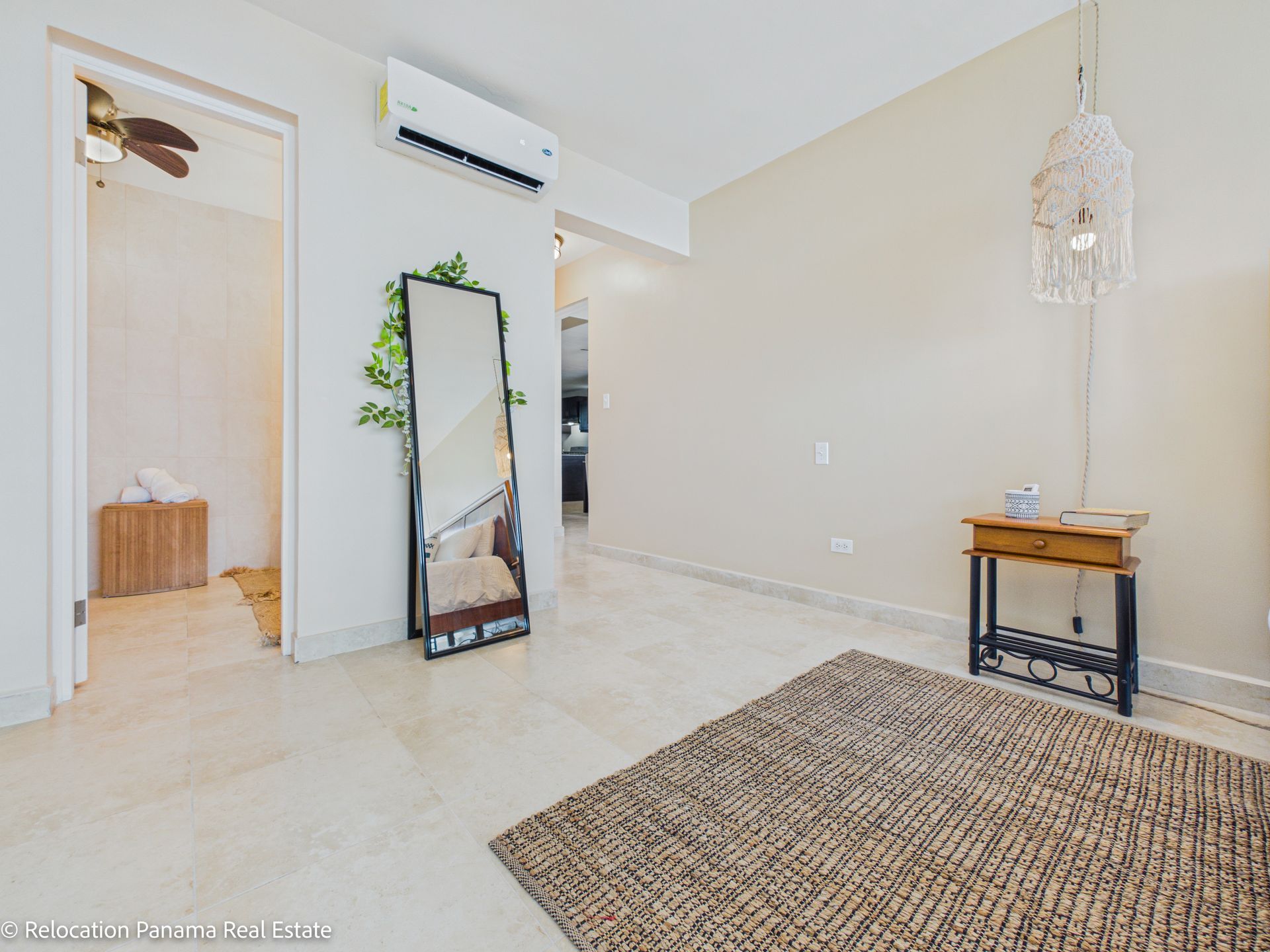 Una habitación luminosa con paredes de color beige, suelo de baldosas claras, una alfombra tejida, un espejo de pie y una pequeña mesa consola de madera.