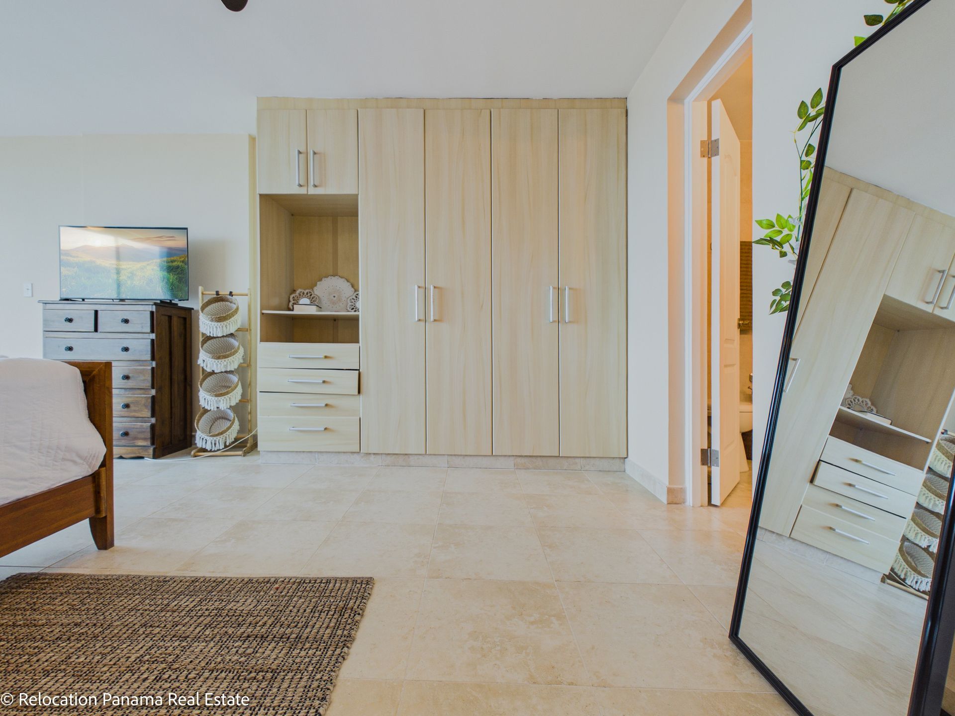 Un dormitorio luminoso y moderno con un armario de madera clara, una cómoda con televisor, un espejo y una alfombra tejida.
