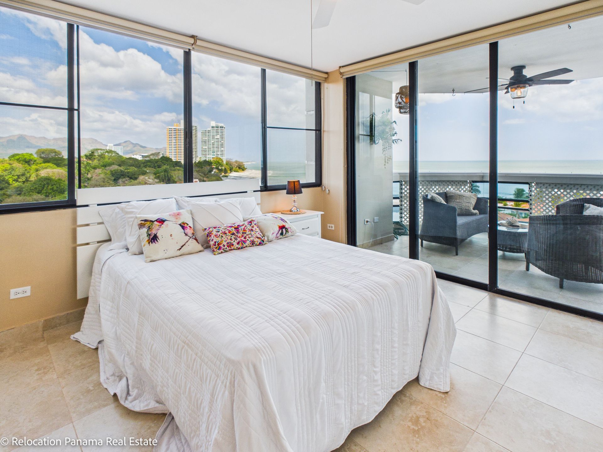 Un dormitorio luminoso y moderno con una cama con ropa de cama blanca, suelos de baldosas y ventanales que van del suelo al techo con vistas al mar.