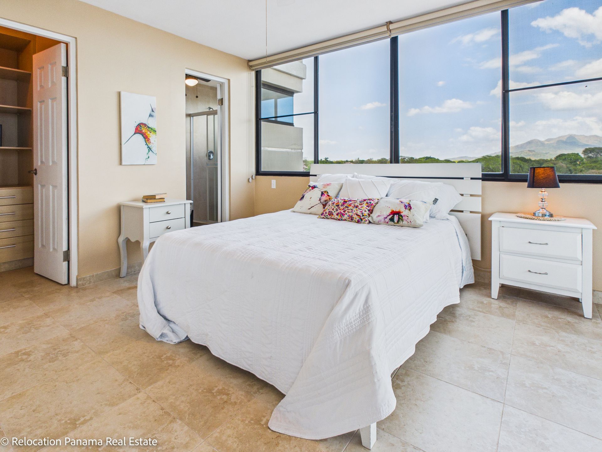 Un dormitorio luminoso con ropa de cama blanca, cabecero, dos mesitas de noche blancas y grandes ventanales con vistas panorámicas al exterior.