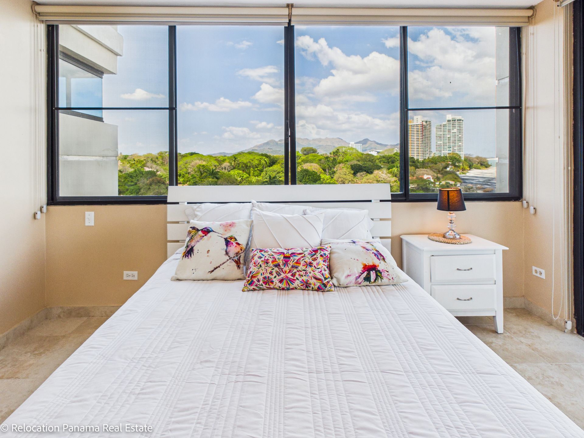 Una cama con ropa de cama blanca, almohadas estampadas y cabecero de madera, situada en el centro, frente a una ventana con vistas a la ciudad.