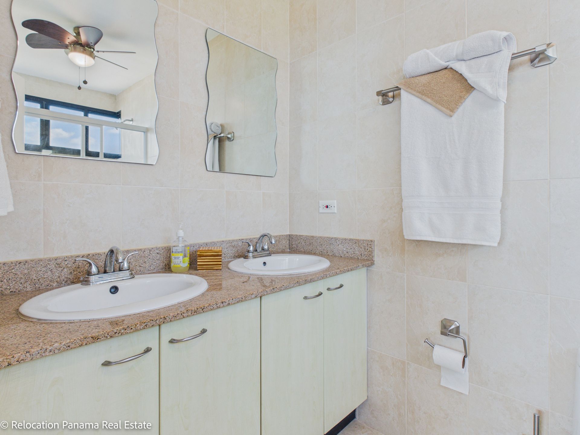 Un moderno mueble de baño con dos lavabos, encimeras moteadas, armarios de color verde pálido, dos espejos y un toallero colgante.