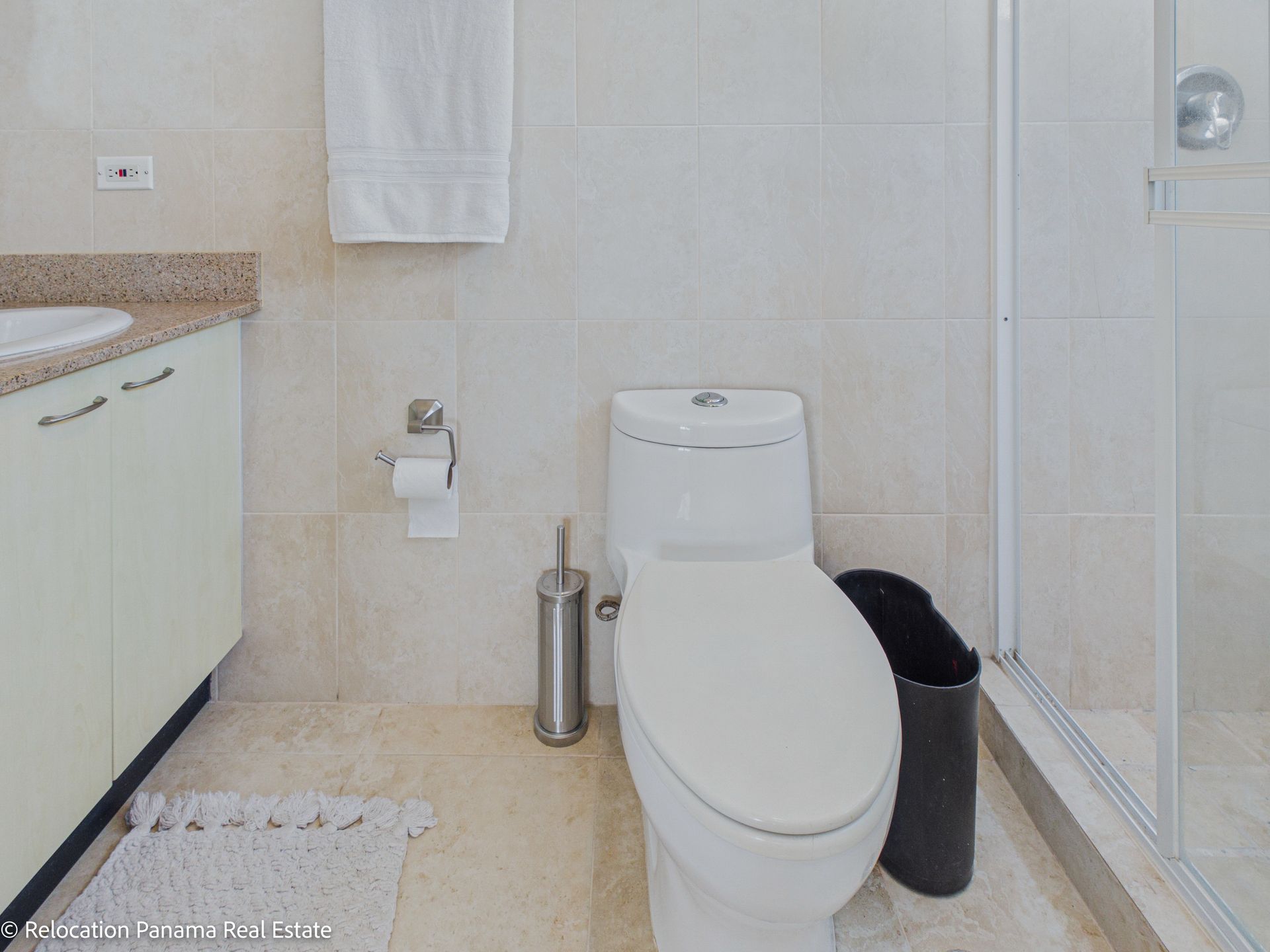 Un inodoro blanco, un desatascador plateado y un cubo de basura negro en un baño con azulejos beige, junto a una ducha con mampara de cristal.