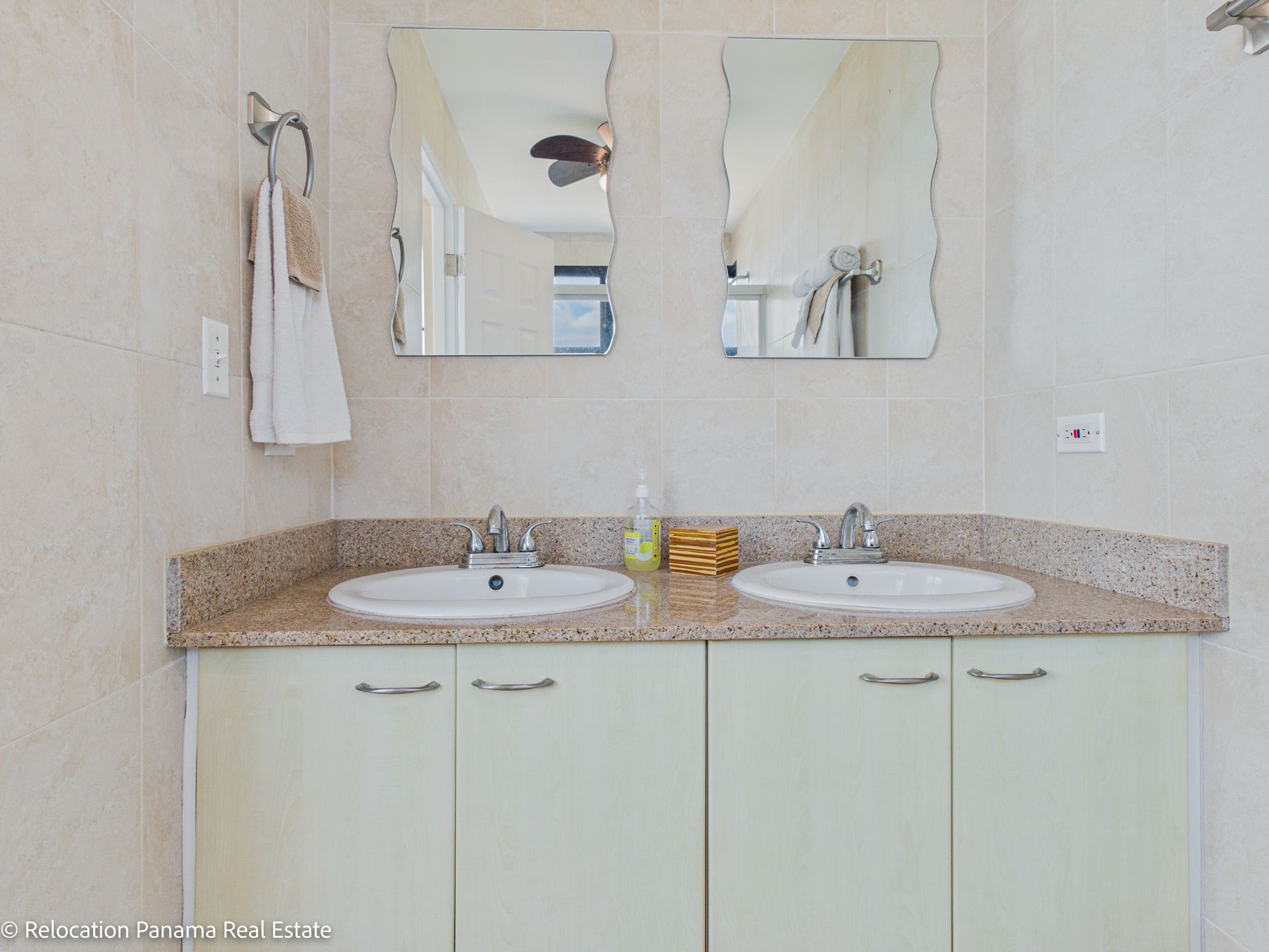 Un tocador de baño con doble lavabo, encimeras de granito moteado beige y armarios de color verde pálido sobre una pared de azulejos claros.